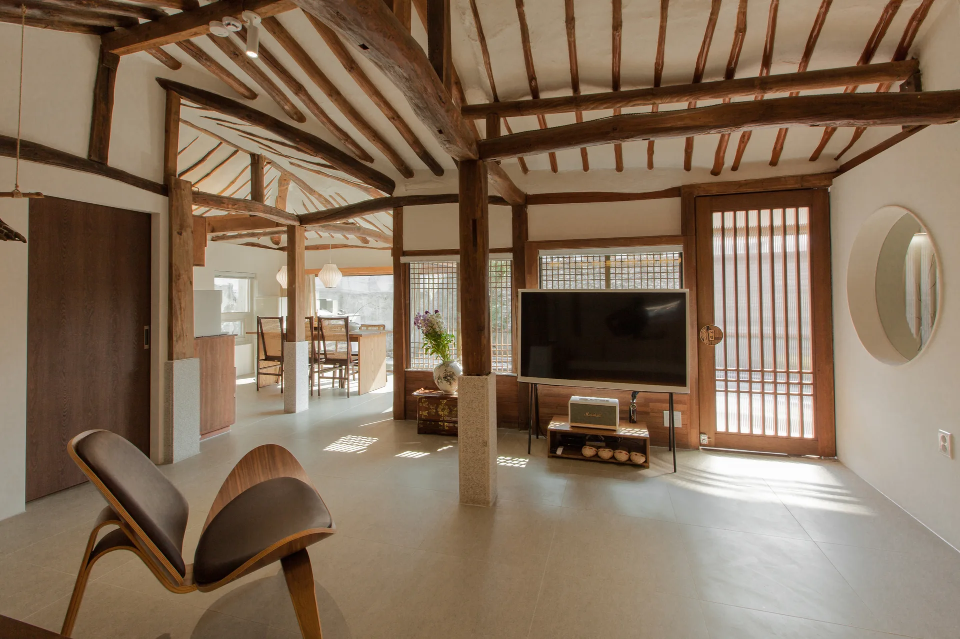 A modern living room features exposed dark wood ceiling beams, a stone pillar, a curved lounge chair, and a wood-framed lattice door.