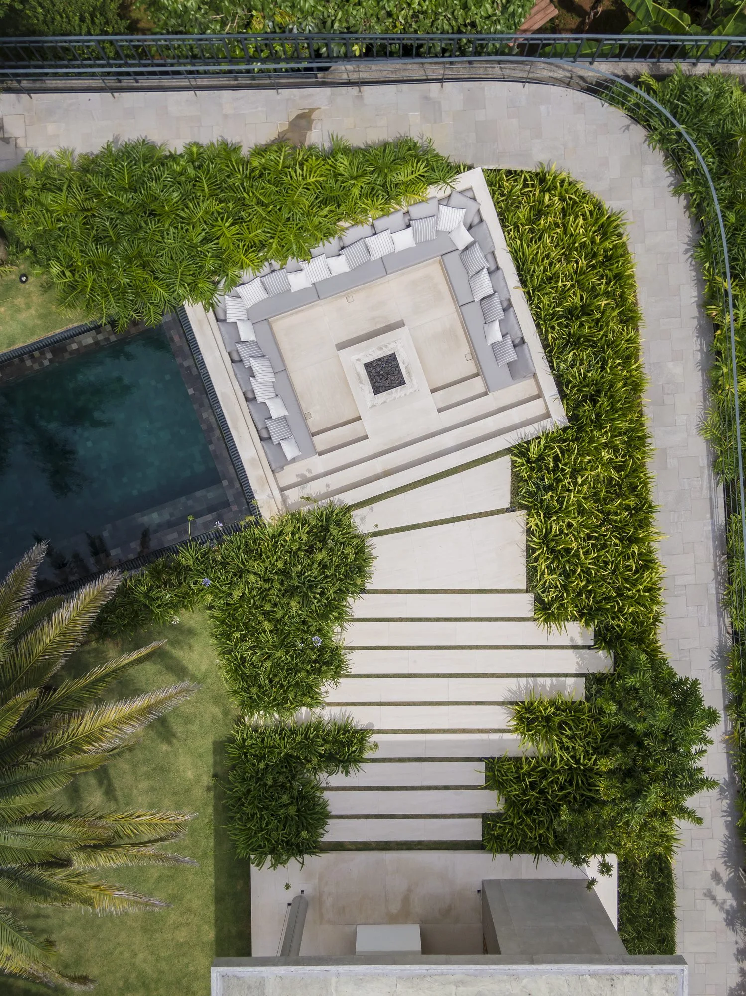 An aerial view of a sunken stone seating area with a central fire pit, tiered steps, and lush green plants next to a pool.