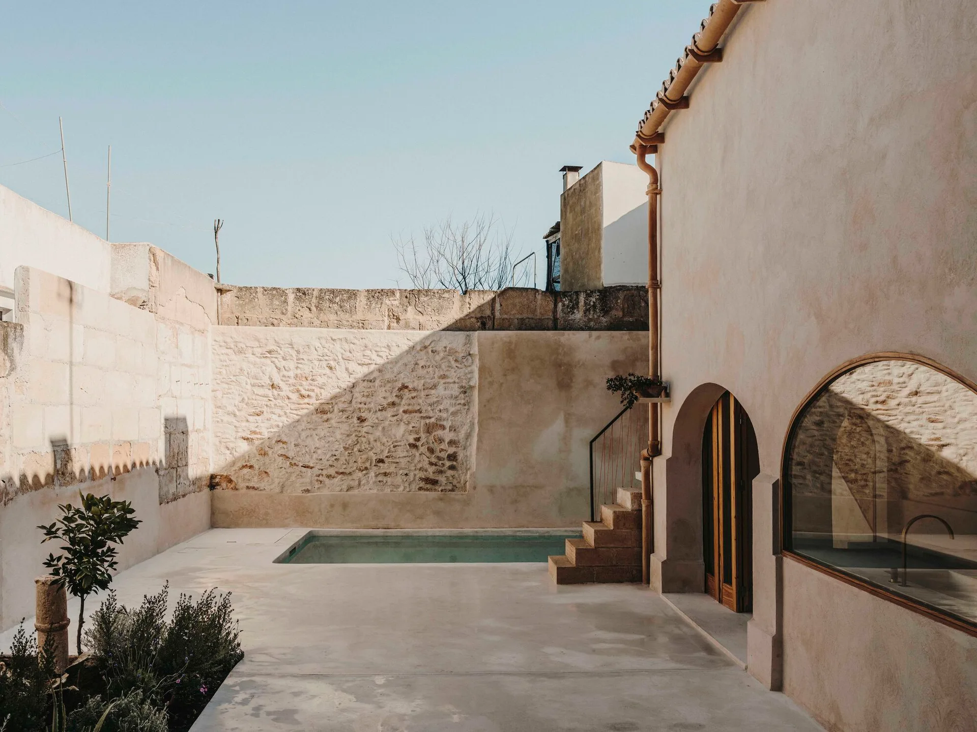 A rectangular swimming pool sits within an enclosed outdoor courtyard featuring warm beige plaster walls, an arched window, and a textured stone wall.