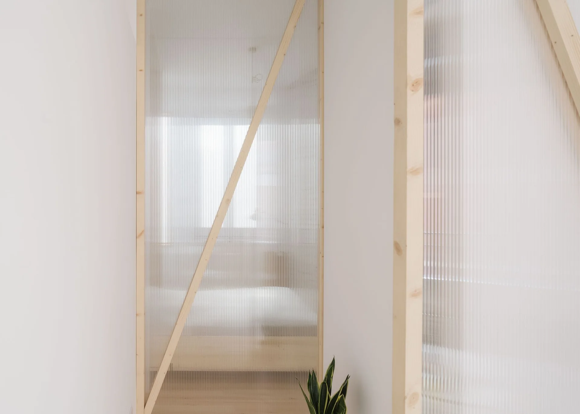A minimalist interior featuring a translucent fluted partition wall framed by light vertical and diagonal wood beams.