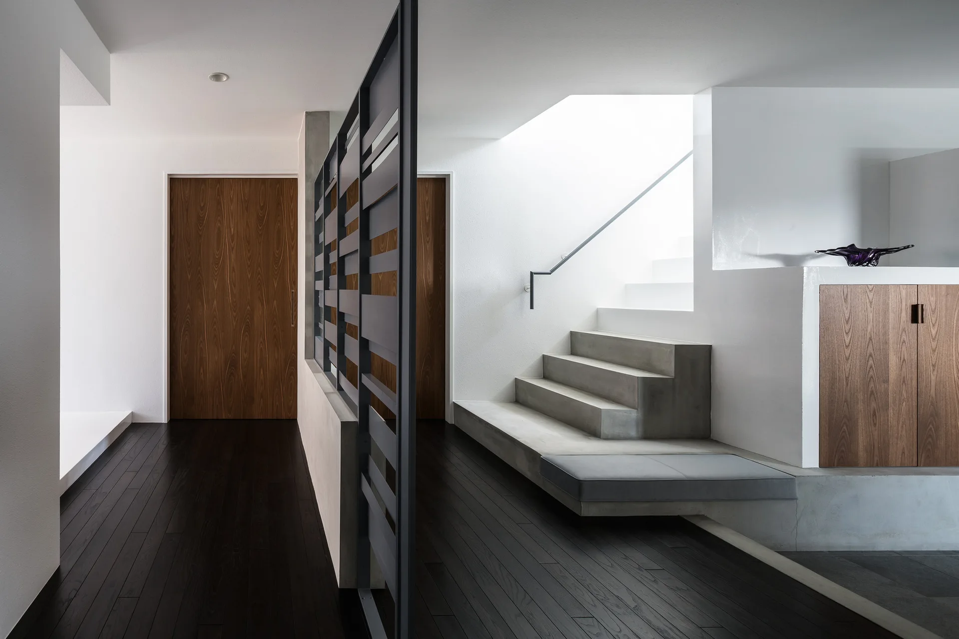 A minimalist modern entryway featuring a concrete staircase, black metal slatted partition, dark hardwood floors, and warm wood doors.