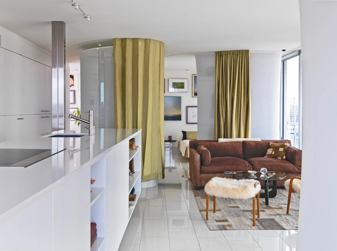 A modern open-plan living room with a brown sofa, furry stools, a glass coffee table, and olive green curtains hanging from a curved ceiling track.
