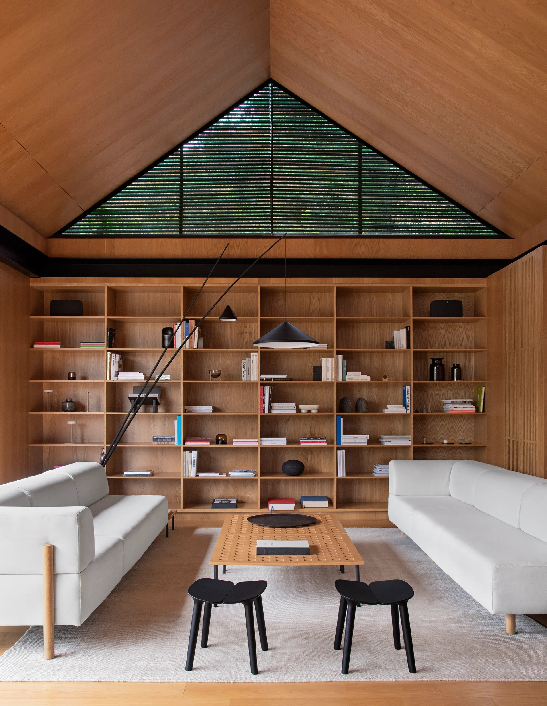 A minimalist living room featuring a vaulted wood ceiling, two white sofas, a central wood coffee table, and a large built-in bookshelf.