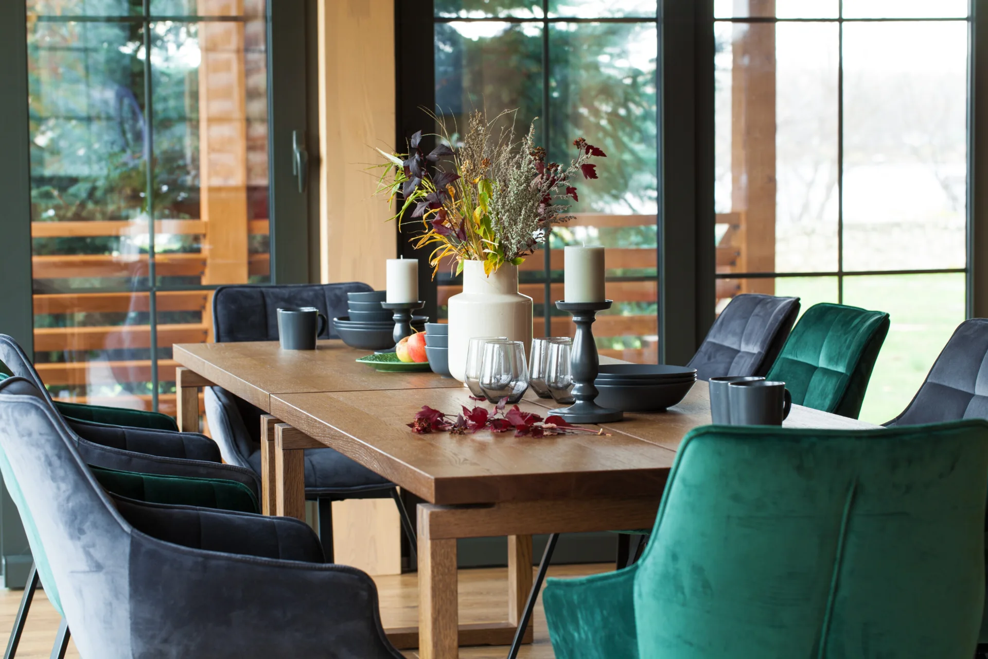 Upholstered velvet chairs in emerald green and charcoal surround a wooden dining table set with ceramic dishes in front of large black-framed windows.