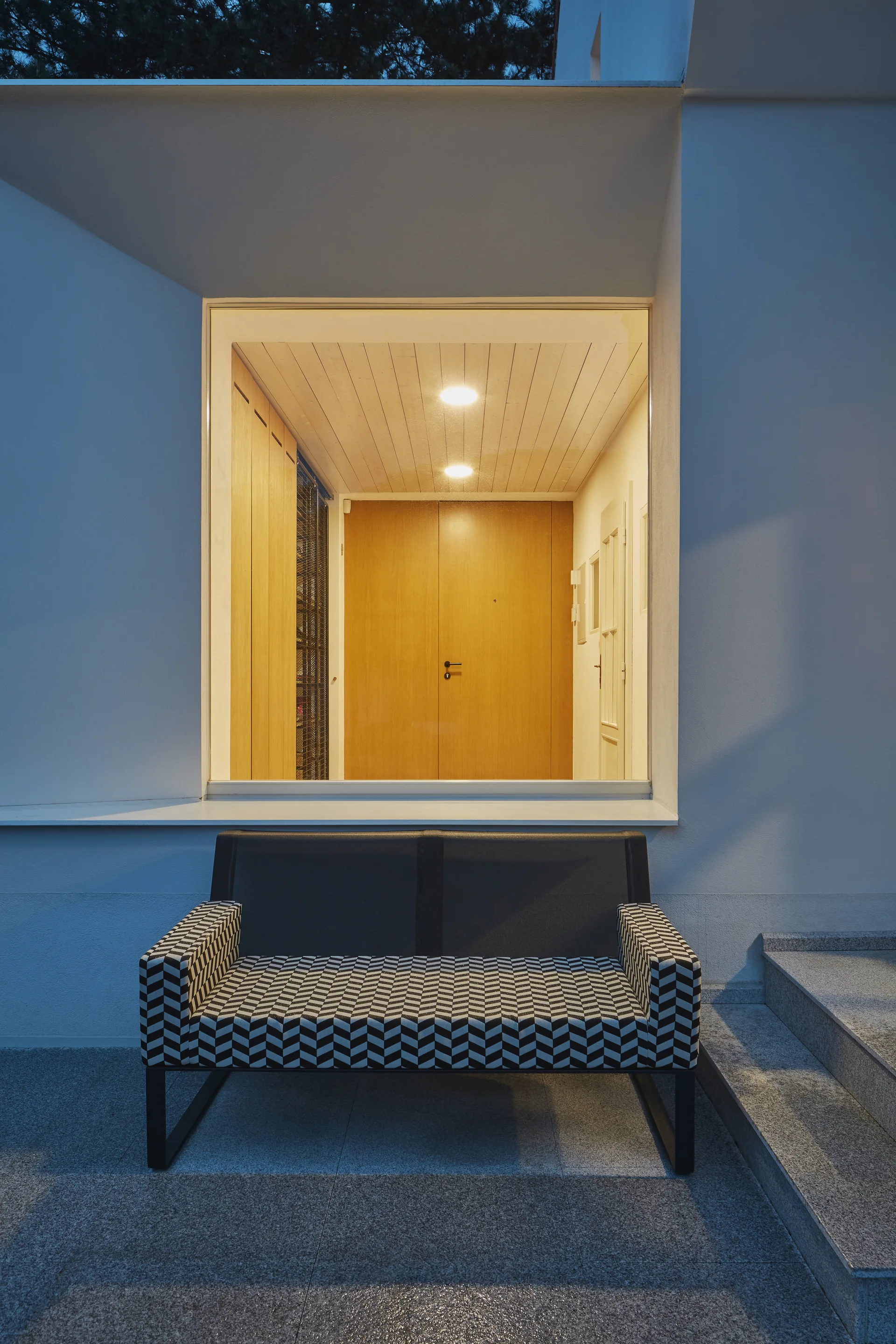 A black and white chevron patterned bench sits on a stone patio outside a large window showing a lit entryway with light wood ceilings.