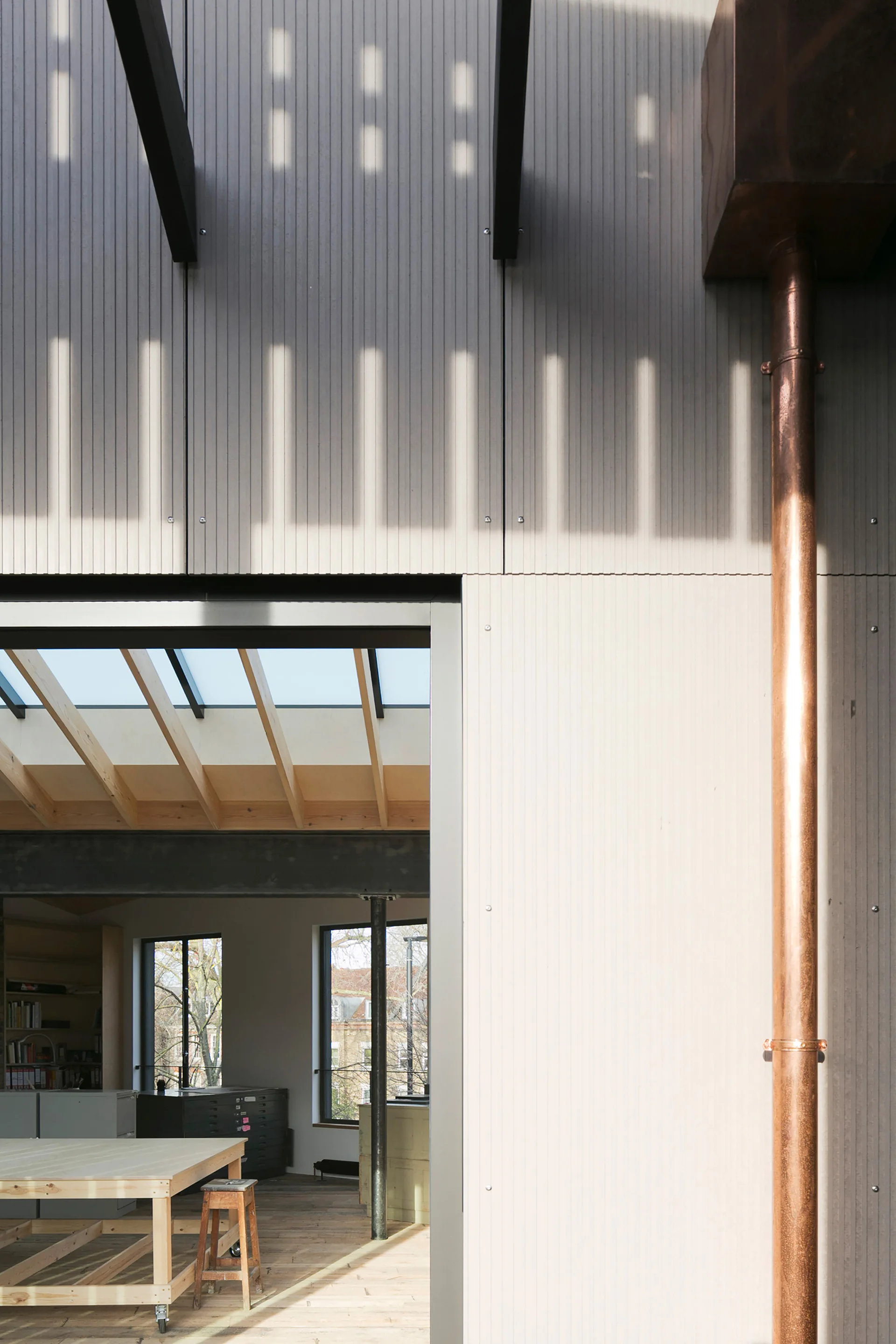 A copper downspout runs down a ribbed grey exterior wall next to an open doorway revealing a bright studio with timber ceiling beams and a skylight.