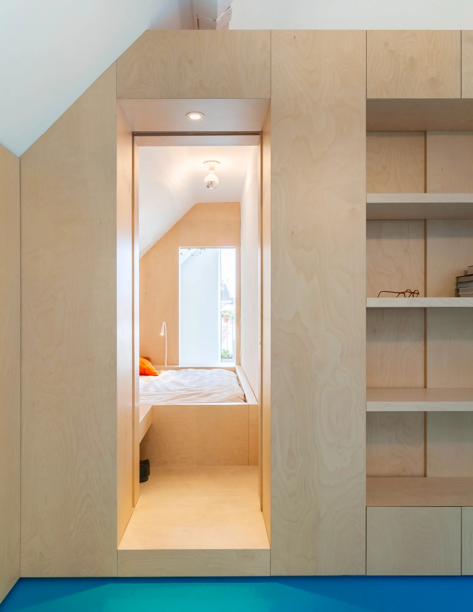 A light plywood storage wall with integrated shelves features a doorway leading into a minimalist attic bedroom nook with a built-in platform bed.