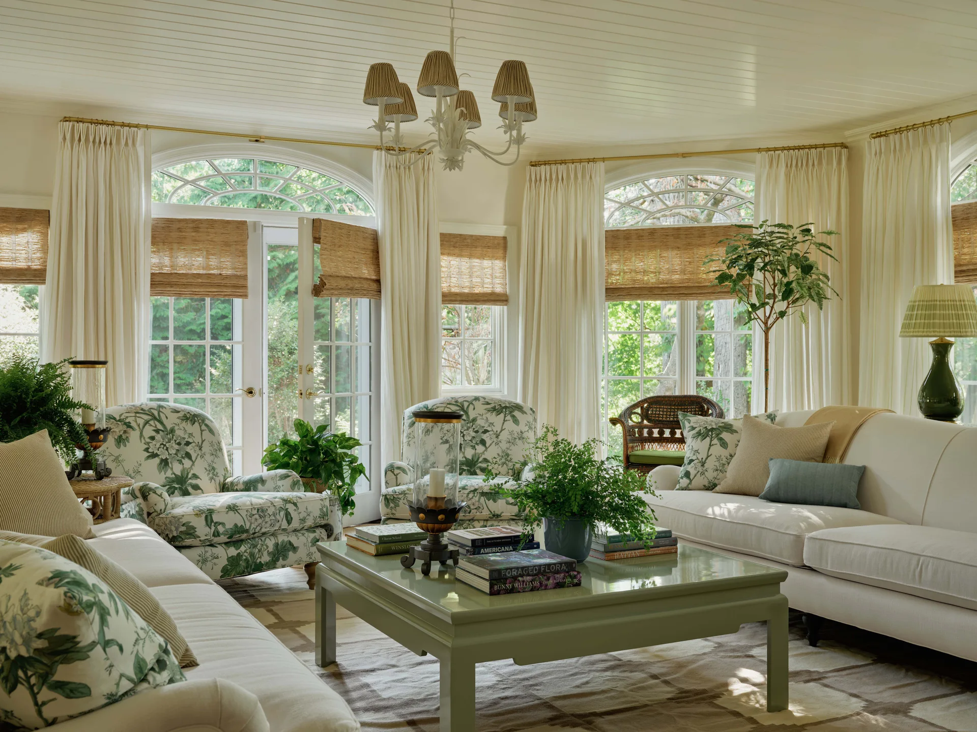 A bright sunroom featuring cream sofas, green floral patterned armchairs, and a sage green coffee table set in front of large arched windows.