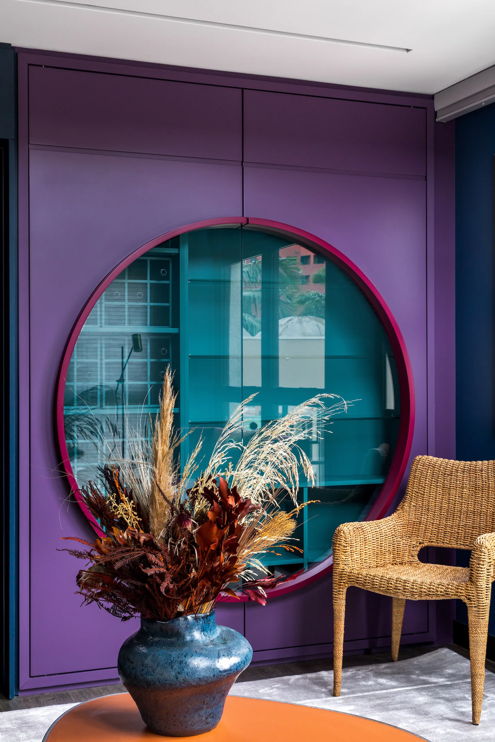 A vibrant purple built-in wall cabinet features a circular glass display opening with teal shelves next to a woven wicker armchair.