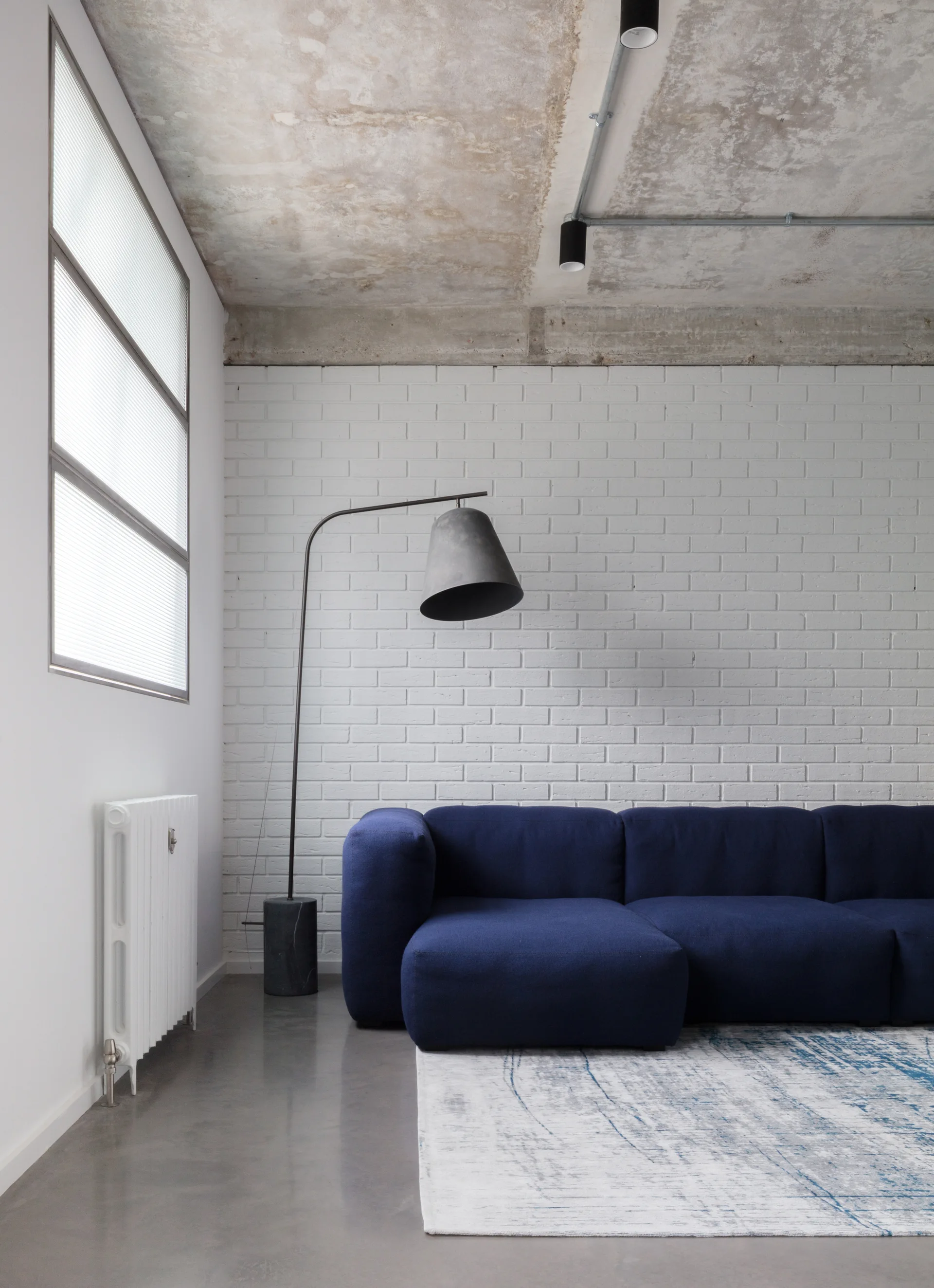A navy blue modular sofa sits on a patterned rug in front of a white painted brick wall and beneath an exposed concrete ceiling.
