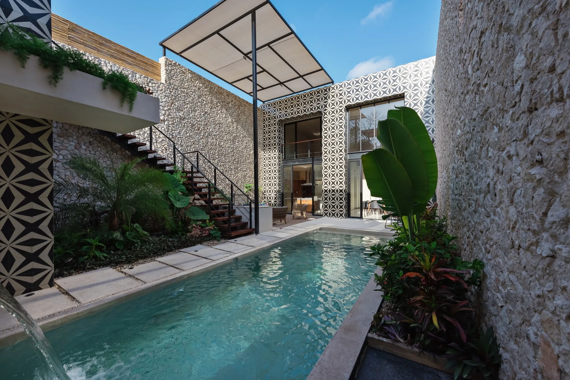 A rectangular swimming pool in an outdoor courtyard flanked by high rough-stone walls, featuring a bold geometric patterned tile accent wall and open stairs.