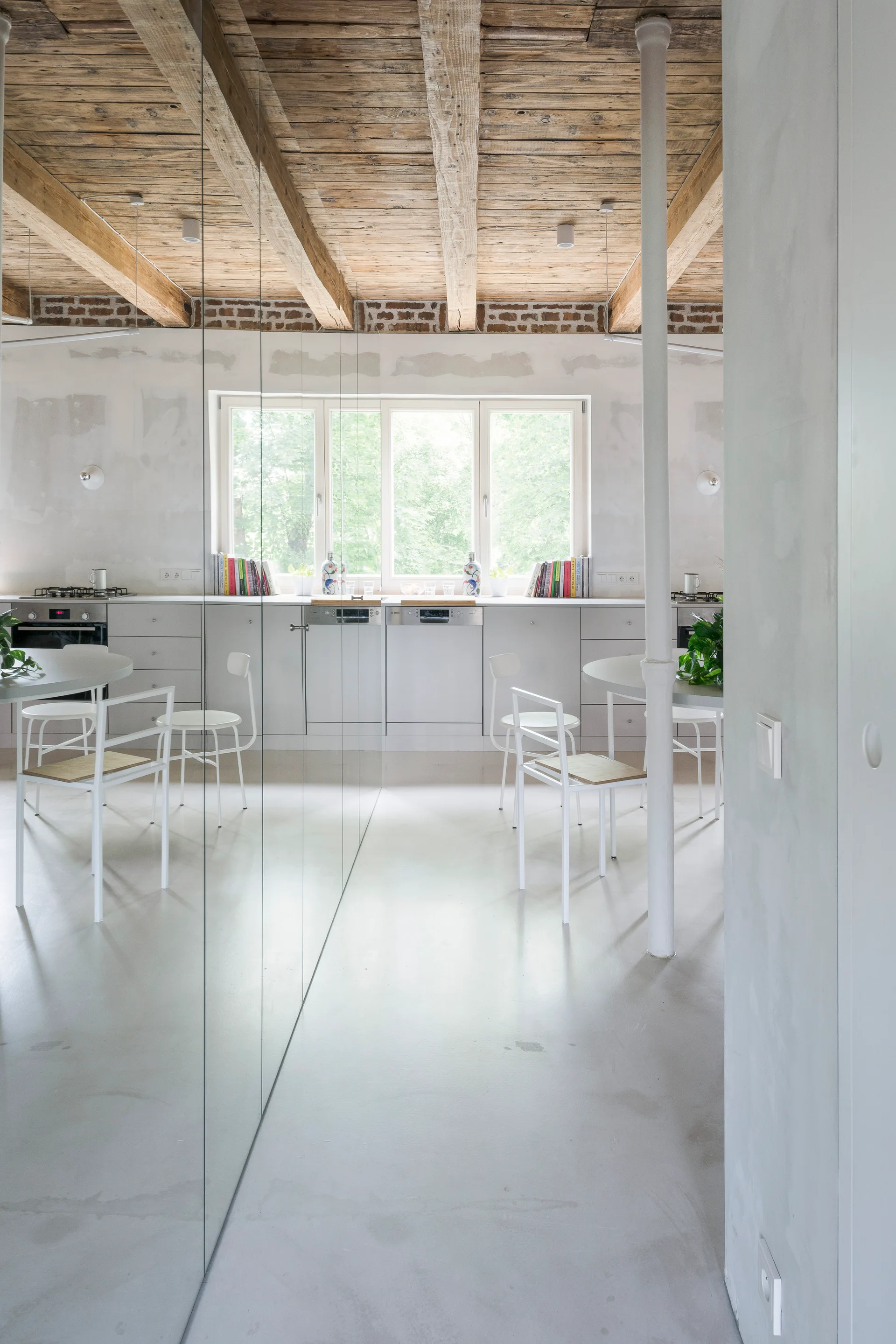 A minimalist kitchen features a long white cabinet run, exposed wood ceiling beams, and a large floor-to-ceiling mirror reflecting a dining table.
