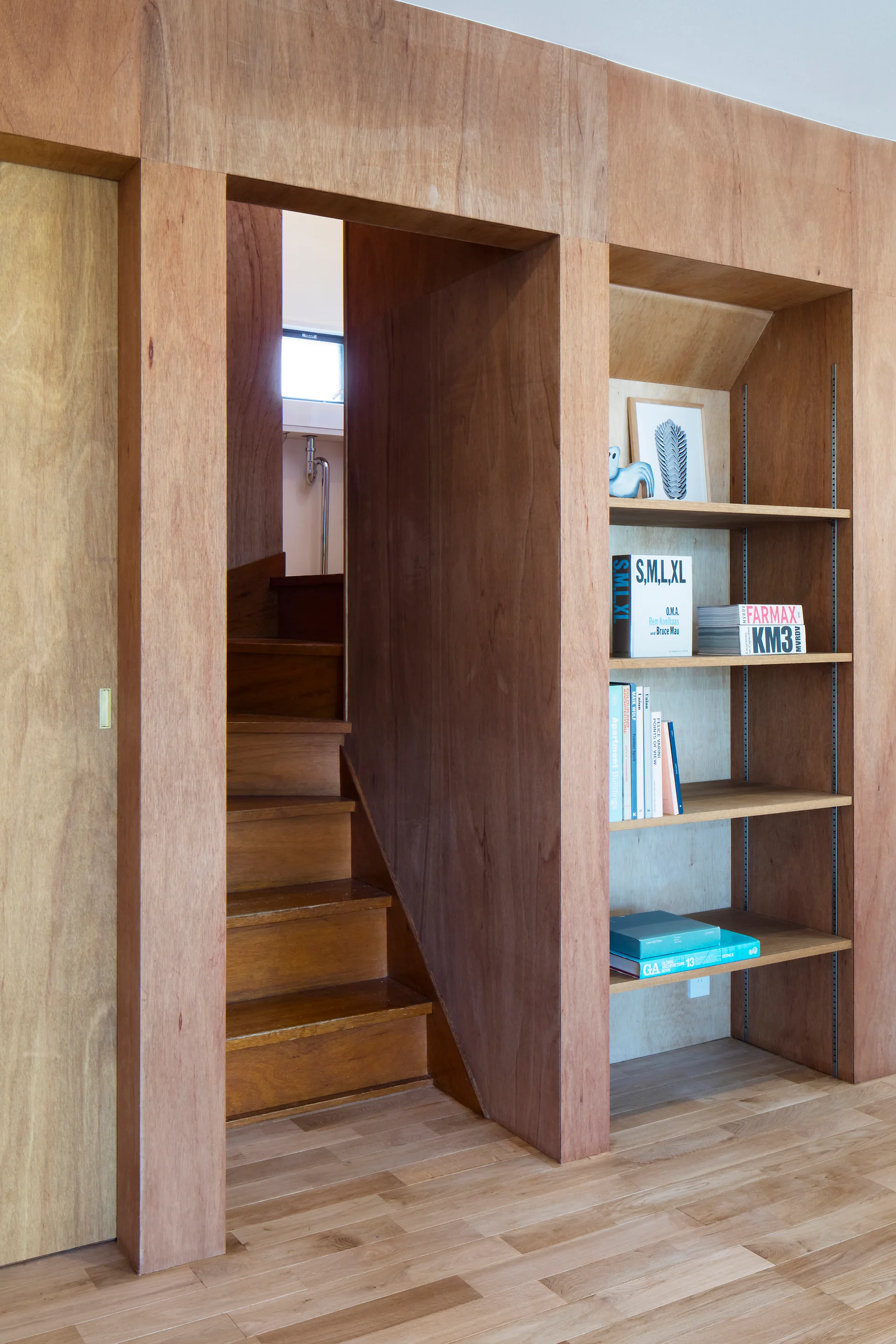 A warm plywood feature wall integrates a central wood staircase entryway, built-in open shelving with books, and a sliding door.