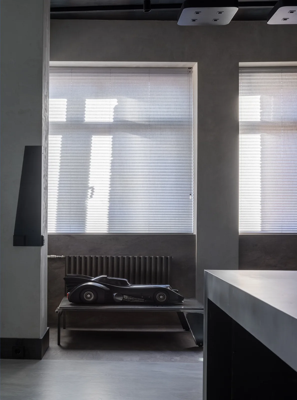 A minimalist grey room with textured concrete walls, a dark metal bench holding a black toy car, and windows with pleated blinds.