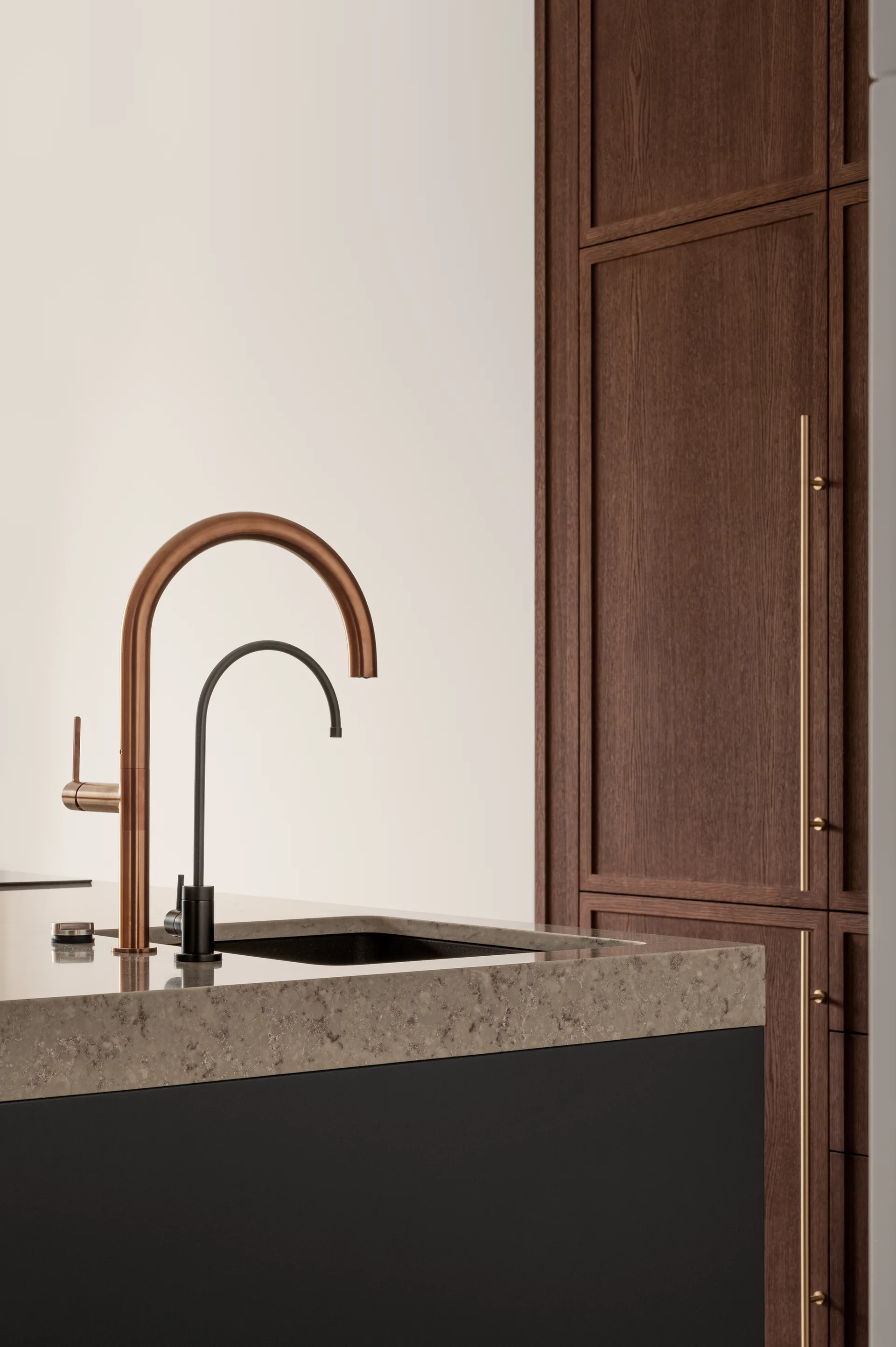 A close-up of a speckled stone kitchen island with copper and black faucets in front of tall dark wood cabinets with long gold pulls.