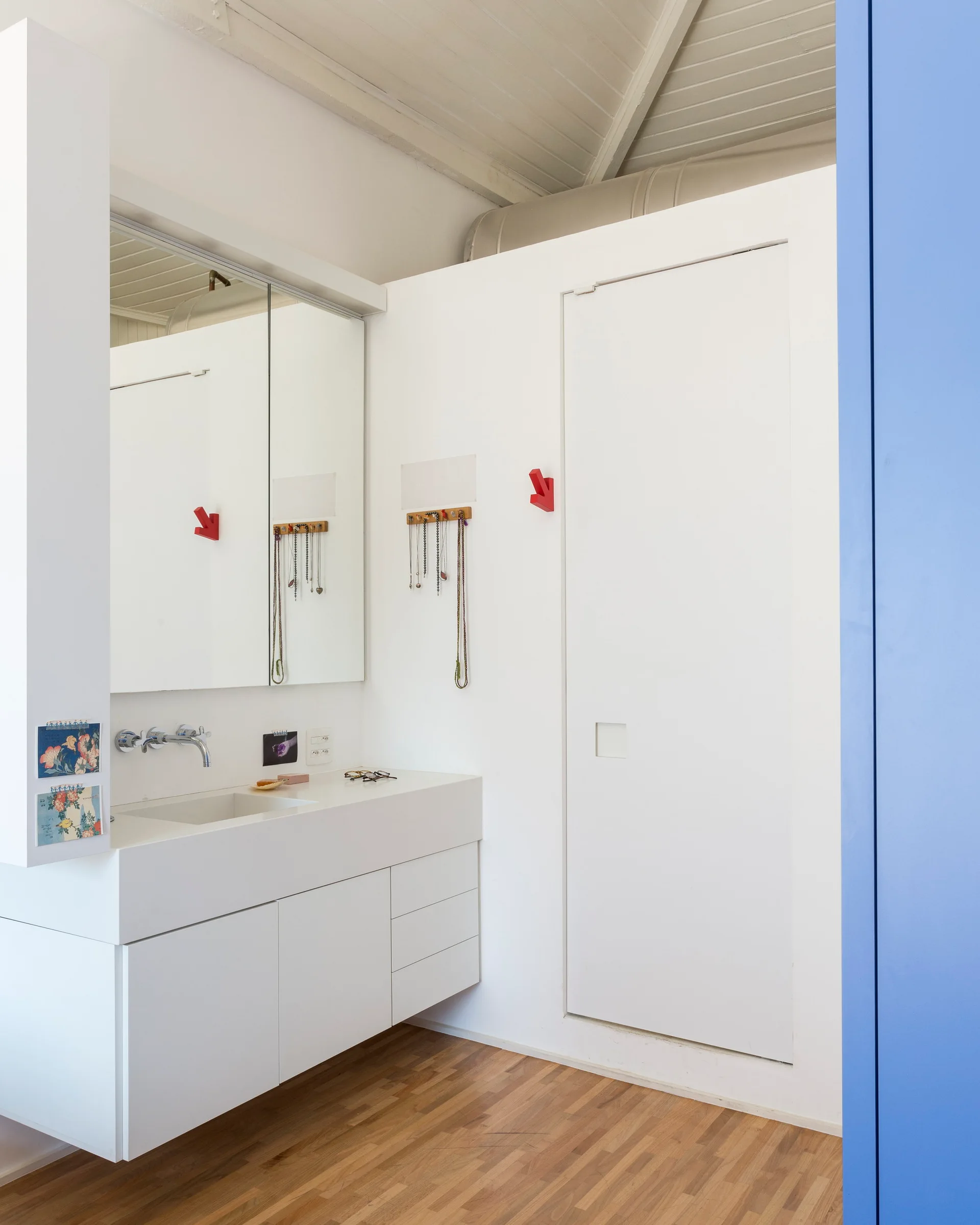 A minimalist white bathroom features a floating white vanity, a large integrated mirror, warm wood floors, and small red arrow-shaped hooks on the wall.
