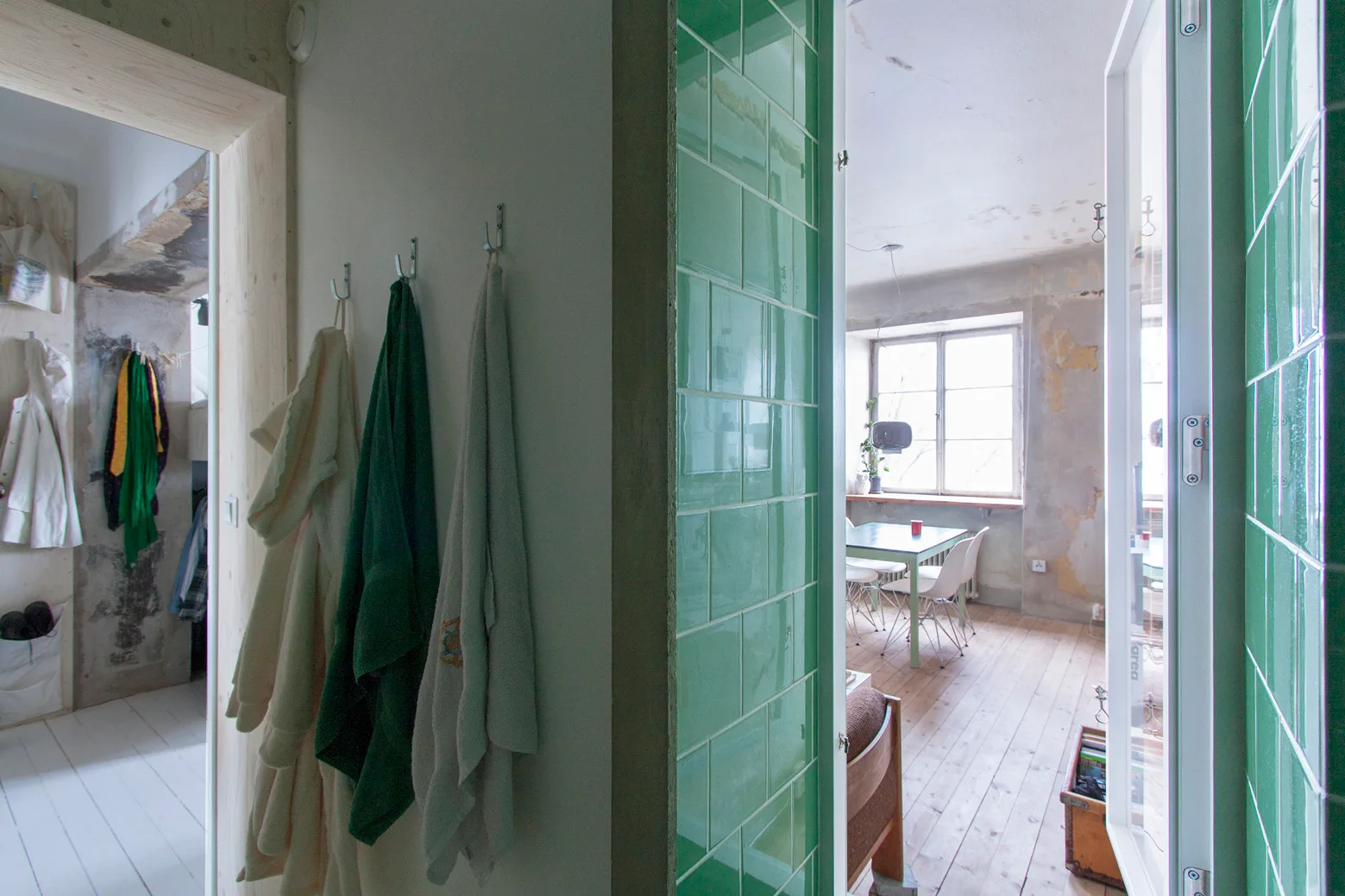 A view through a doorway framed by glossy green tiles looking into a room with raw plaster walls and light wood floors.