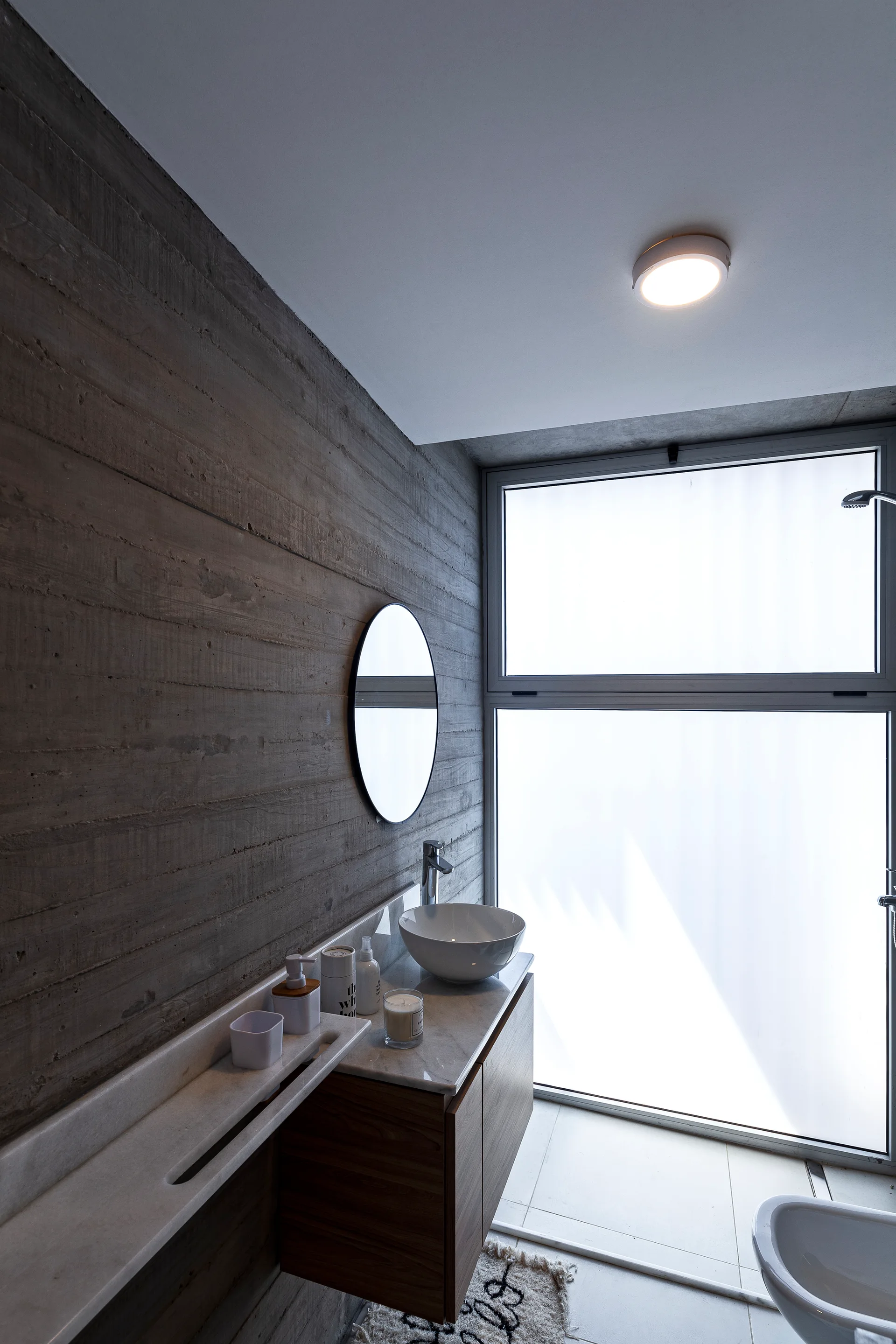 A modern bathroom with a textured board-formed concrete wall, a round mirror, a floating wood vanity, and a large frosted glass window.