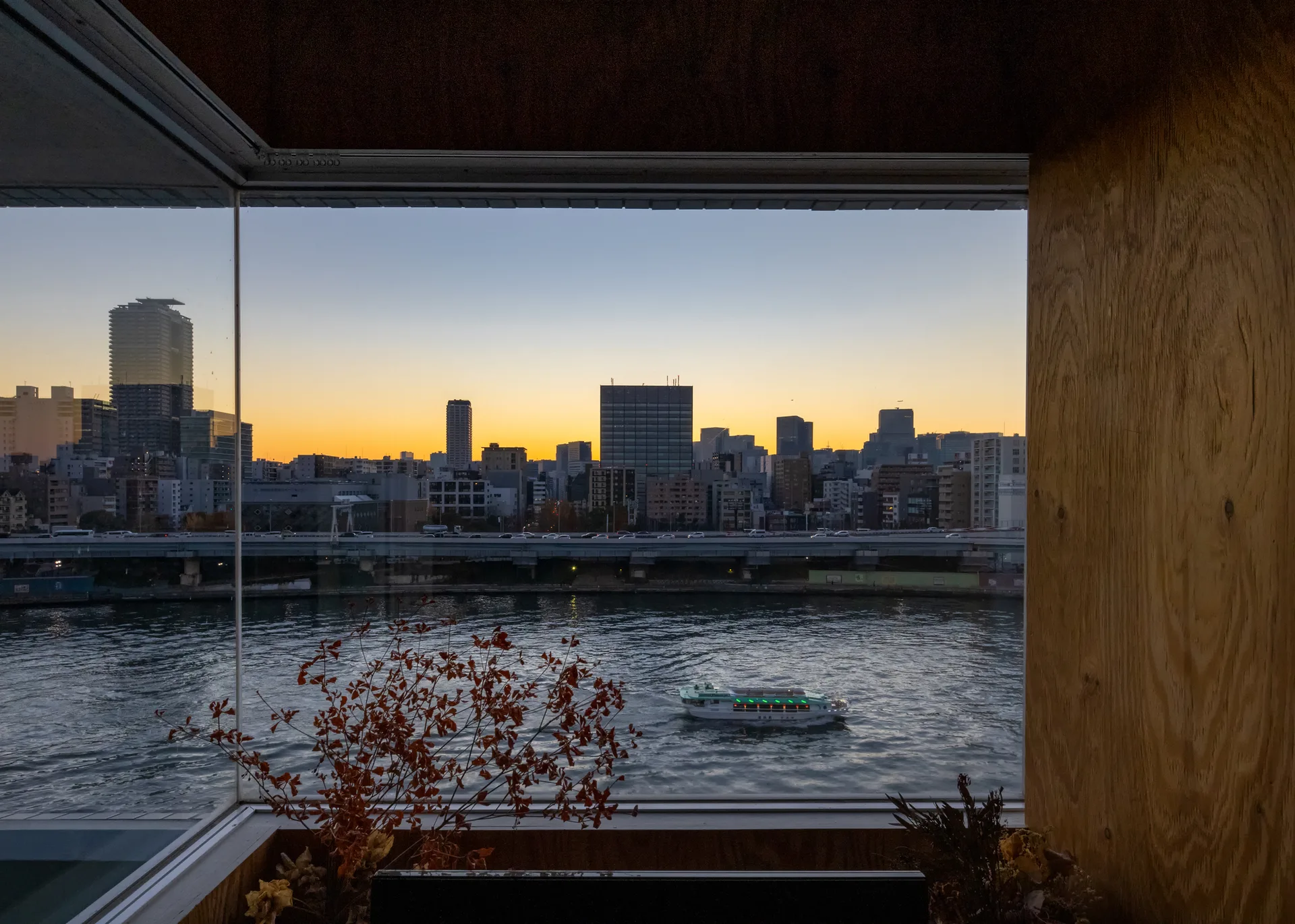 A large window framed in light-toned plywood looks out onto a river and city skyline during a golden sunset.