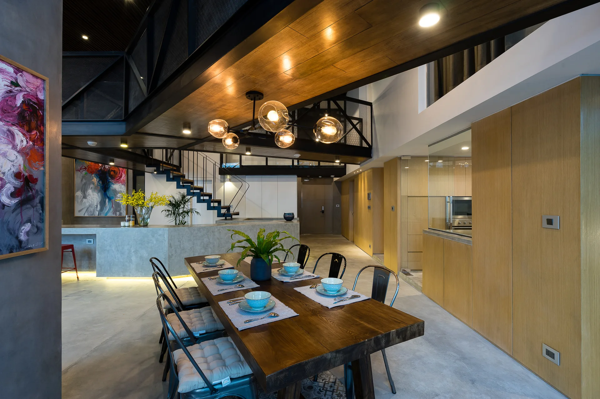 An industrial dining room features a wooden table with metal chairs, a globe pendant light, and a mezzanine structure overhead.