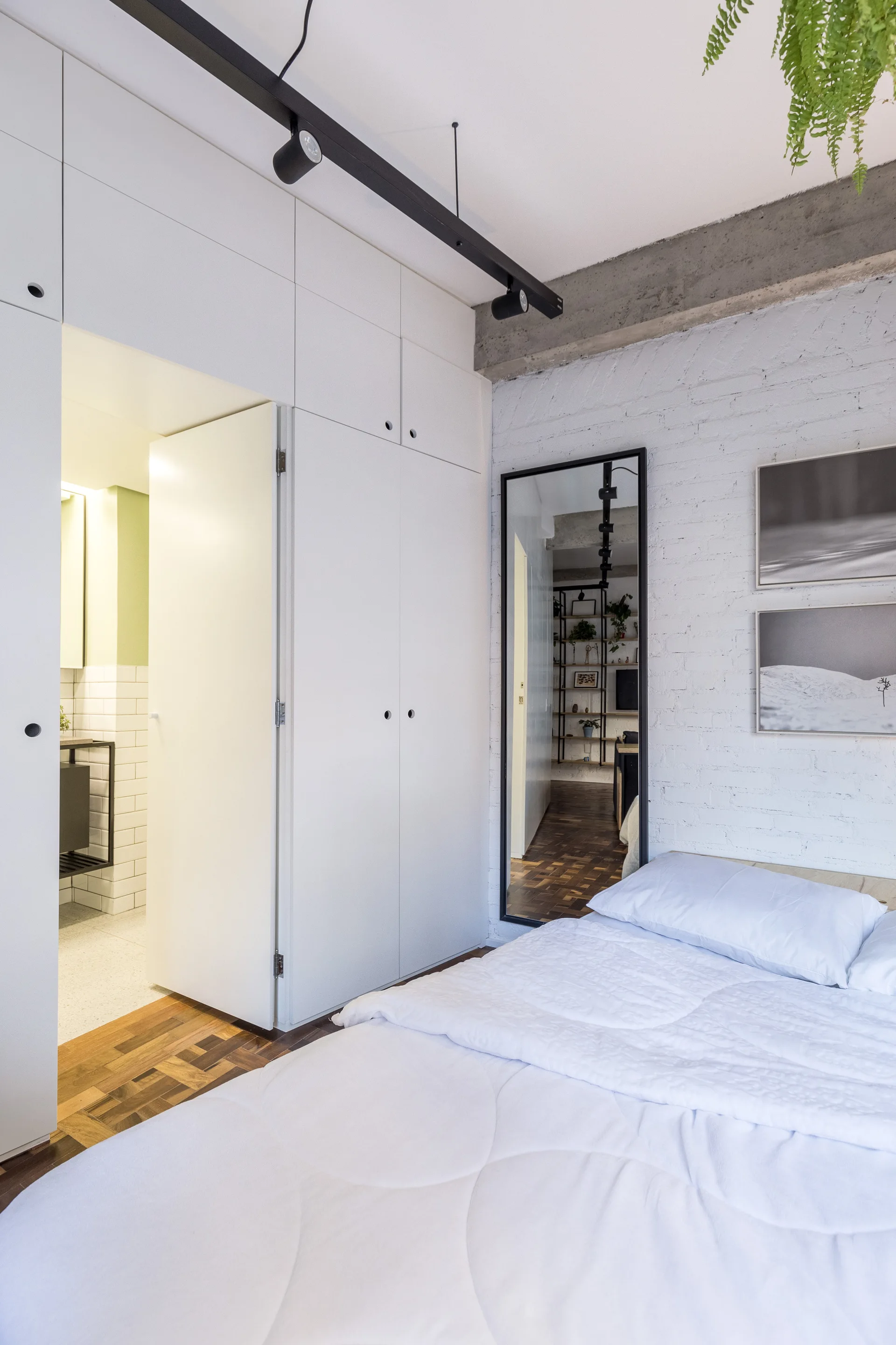 A modern minimalist bedroom featuring white bedding, built-in wardrobes, a tall floor mirror, and a white-painted brick wall under a concrete beam.