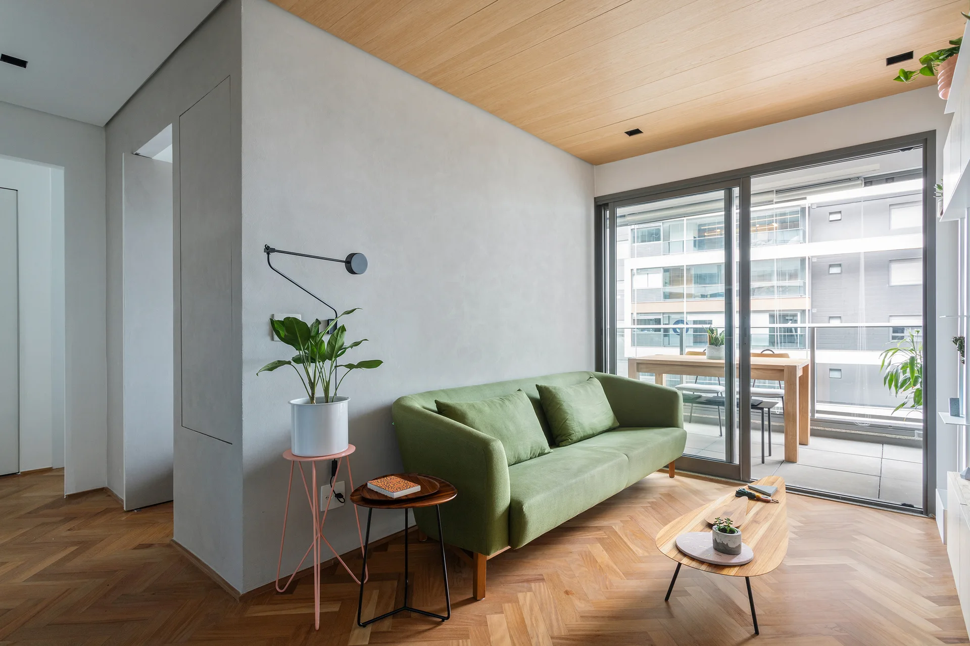 A modern living room features a green sofa on herringbone wood floors beneath a light wood paneled ceiling next to sliding glass doors.