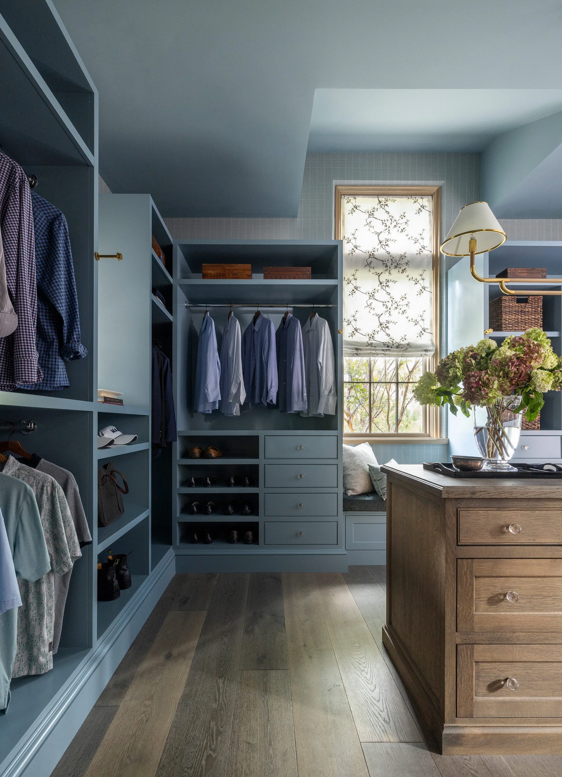 A walk-in closet features slate blue built-in cabinetry, a central wooden storage island, hardwood floors, and a window with a patterned roman shade.
