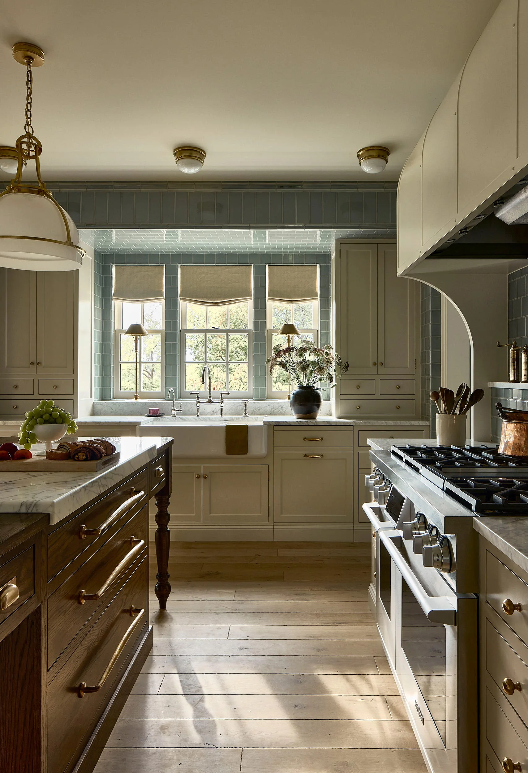 A transitional kitchen features a beige-painted cabinet wall, a blue subway tiled backsplash around triple windows, and a wood island with marble countertops.