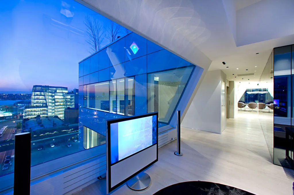 A modern high-rise interior features an angled glass wall overlooking a city skyline at dusk, light wood floors, and a freestanding television.