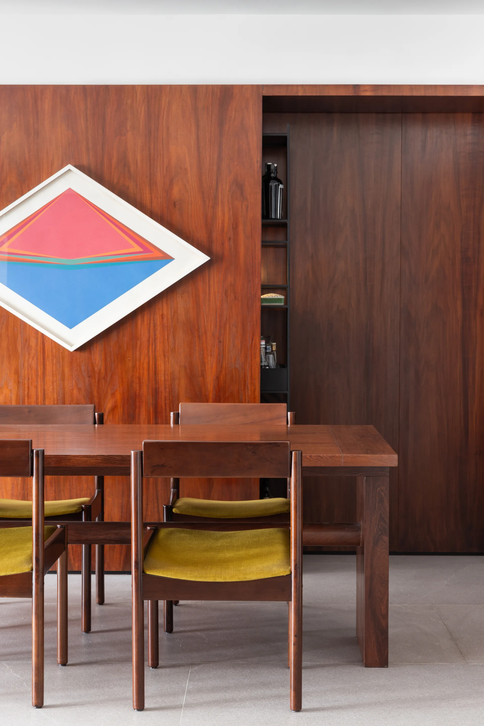 A wooden dining table and chairs with yellow cushions stand in front of a warm wood paneled wall featuring a diamond-shaped abstract painting.