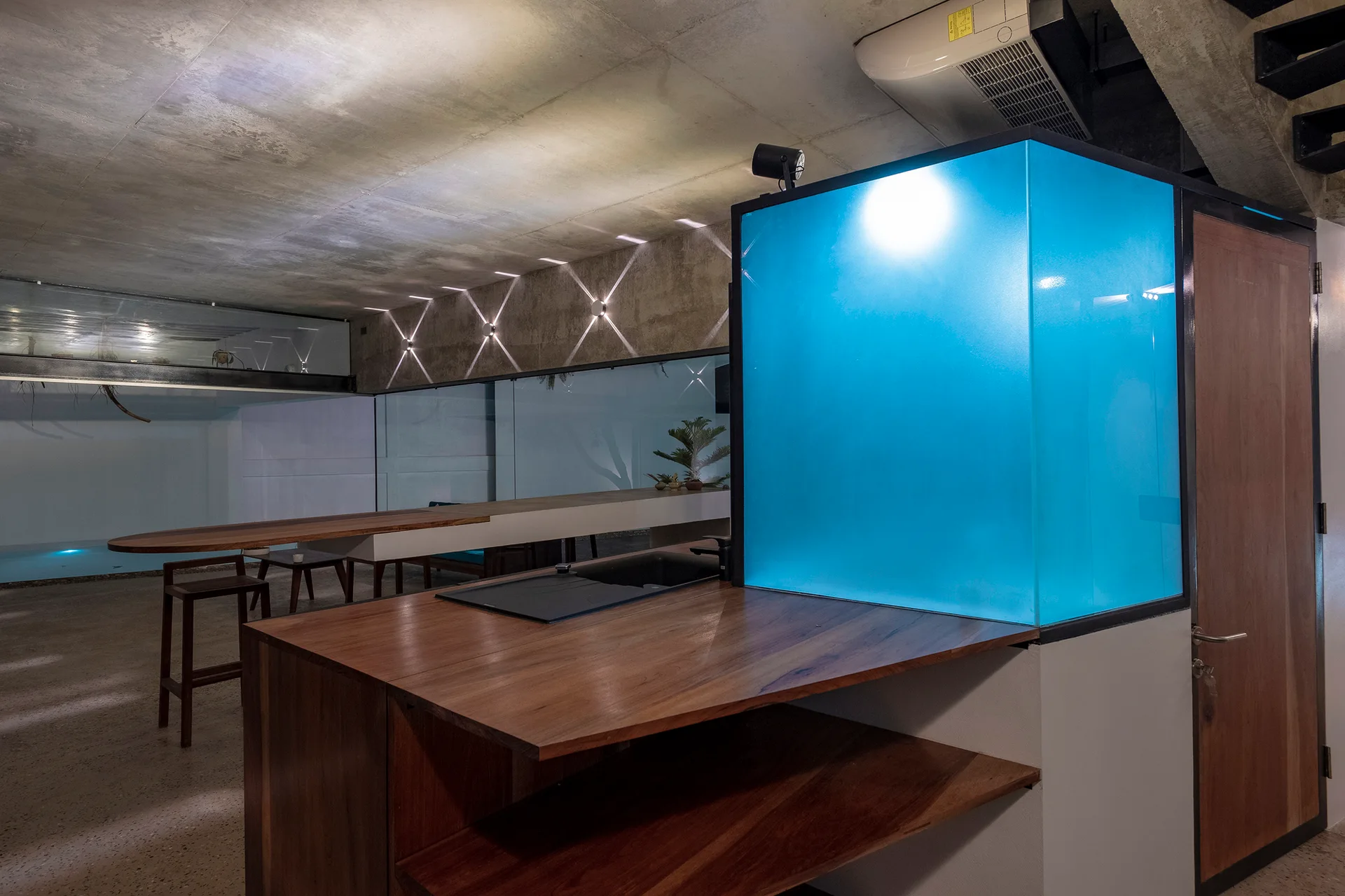 A modern industrial kitchen features a wooden island with a sink next to a glowing blue cube under an exposed concrete ceiling.