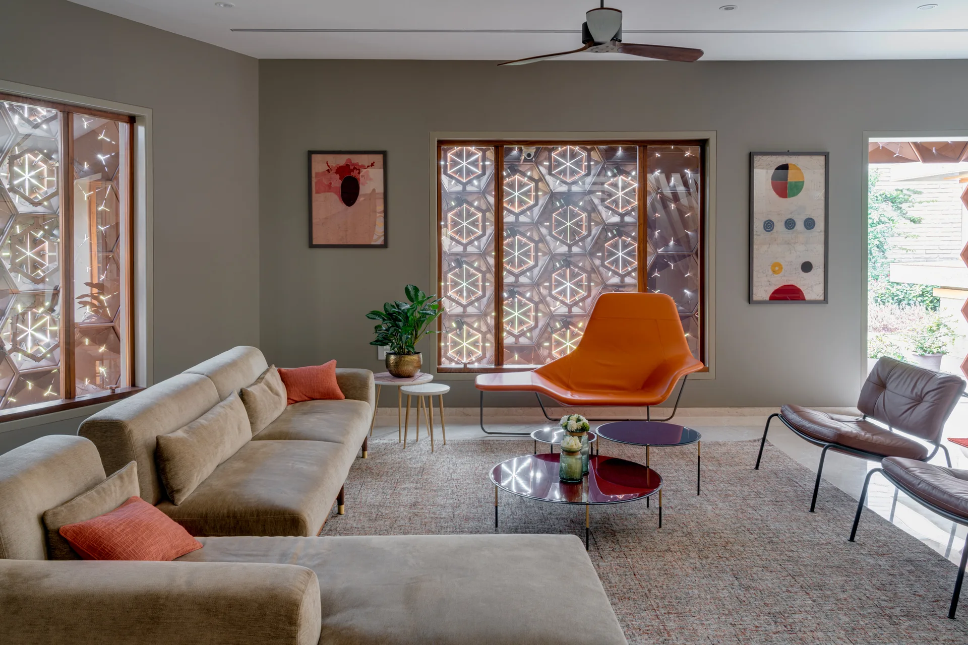 A contemporary living room featuring a tan sectional, an orange lounge chair, and large windows with hexagonal geometric screens.