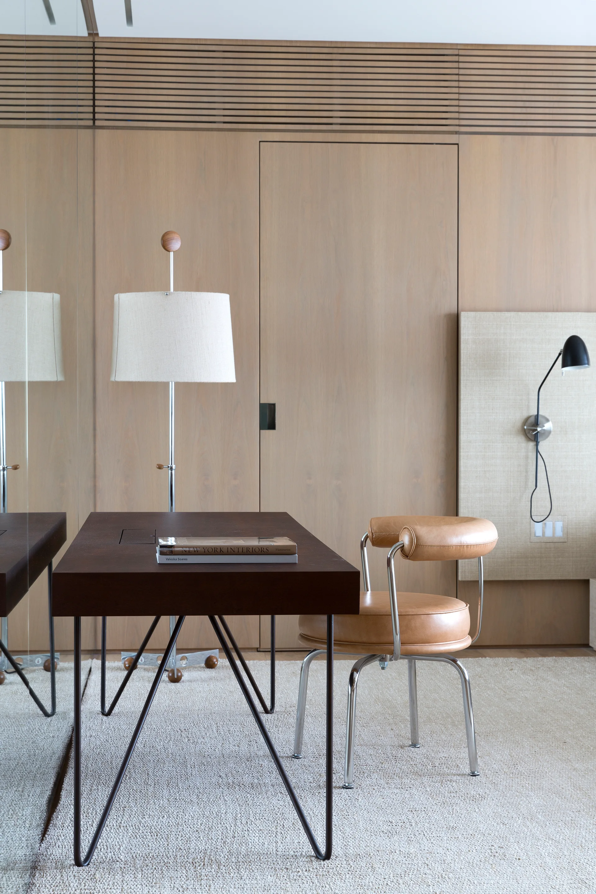 A home office features a dark wood desk and a tan leather chair set against a light oak wood-paneled wall next to a large mirror.
