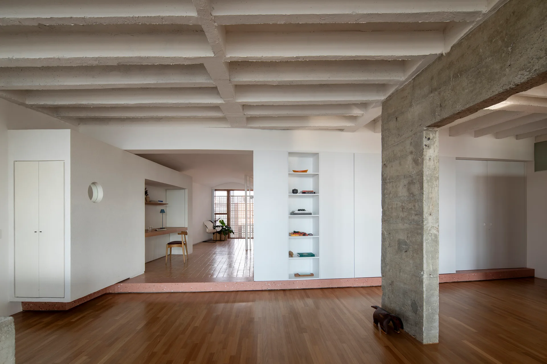 Minimalist interior showing a raw concrete column, an exposed waffle ceiling, wood floors, and a raised study area with terracotta tile flooring.