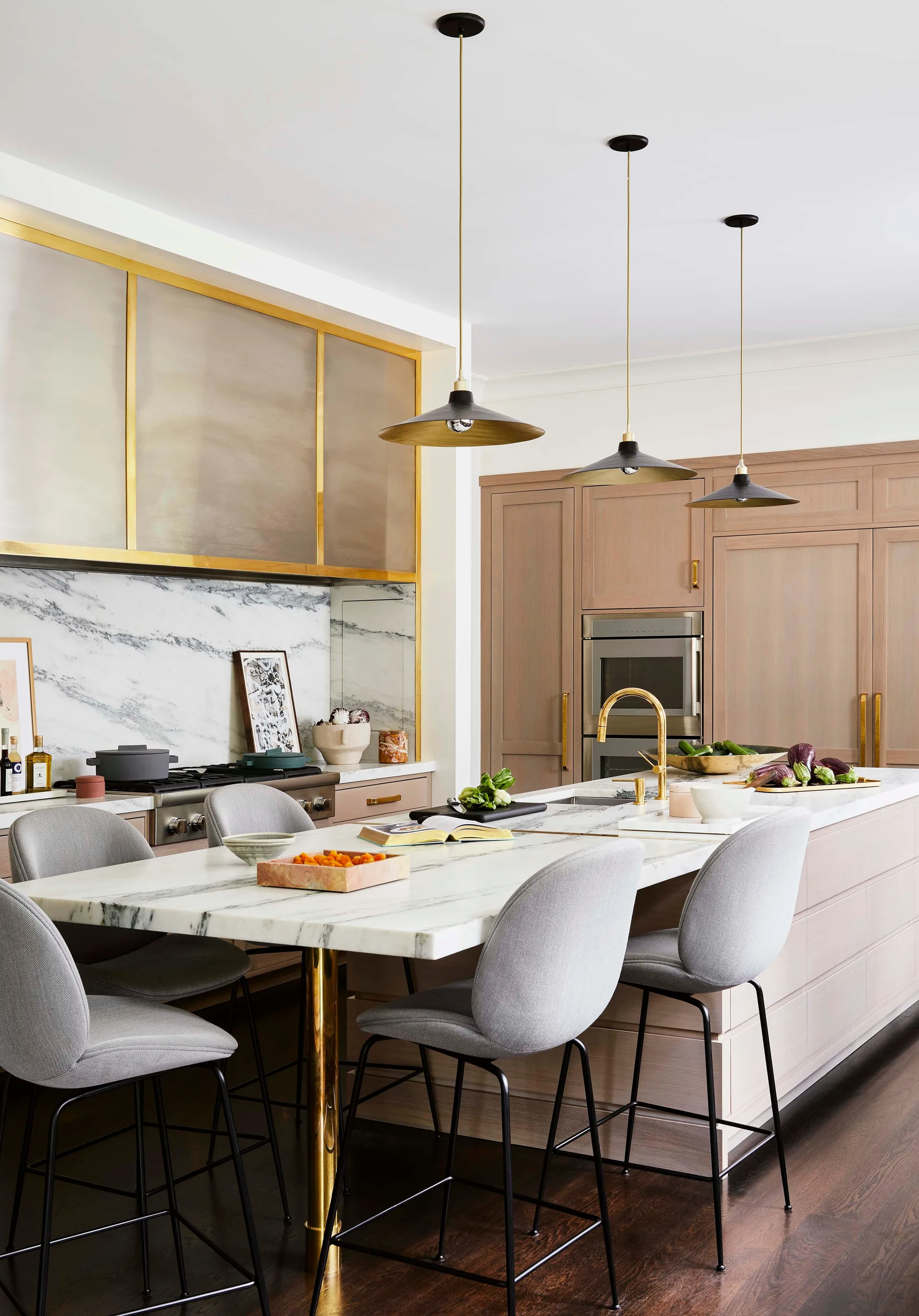 A modern kitchen features a marble topped island with grey stools, brass accented wood cabinetry, and three black pendant lights.