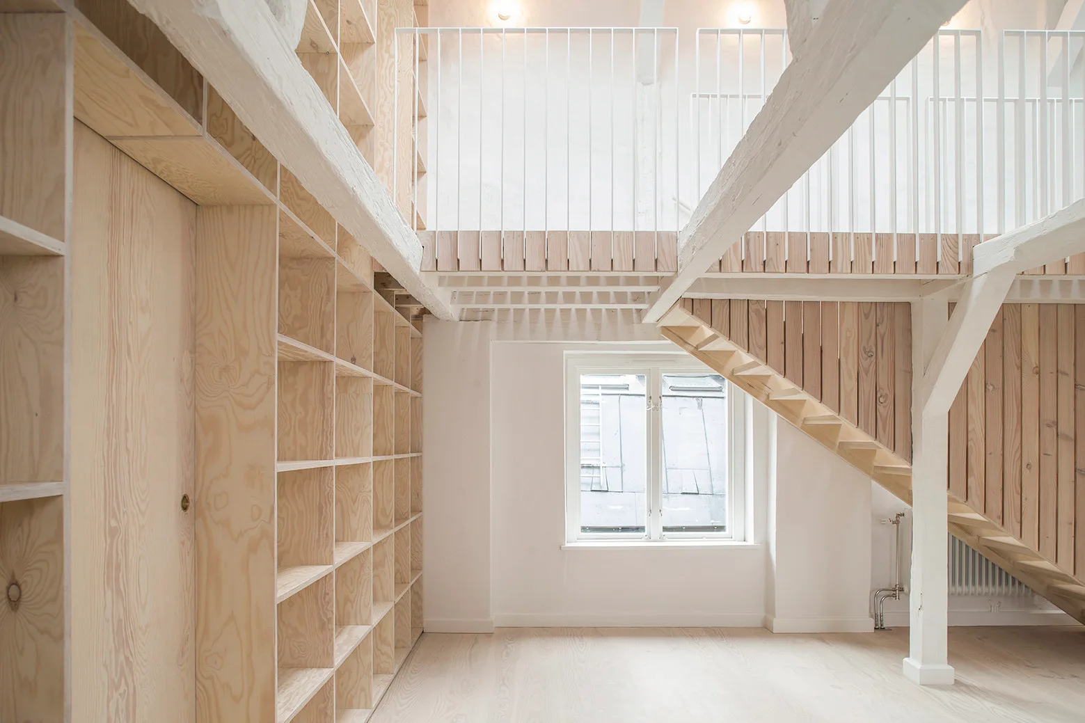 A bright loft interior featuring a tall plywood shelving unit, a wooden staircase leading to a mezzanine, and white painted structural beams.