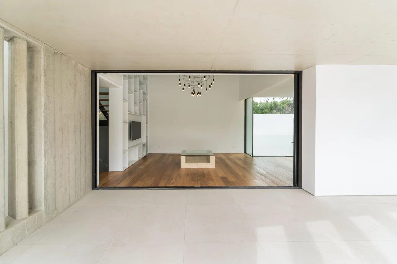 A view through a black-framed opening into a minimalist living room with warm wood floors, white built-in shelves, and a modern pendant light.