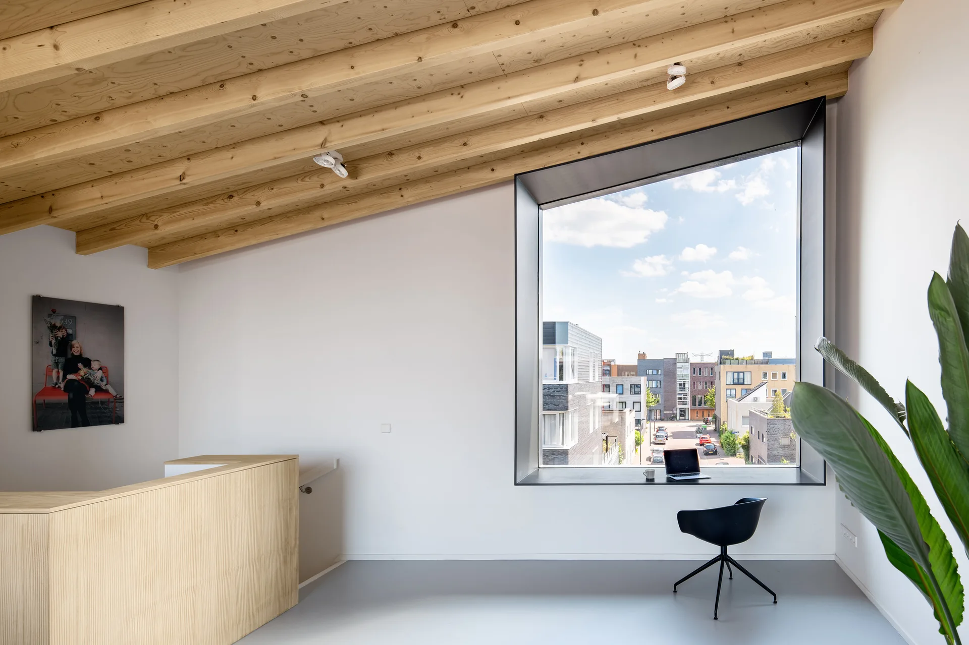 A minimalist workspace features an angled wood-beam ceiling, white walls, and a large deep-set square window used as a desk with a black swivel chair.