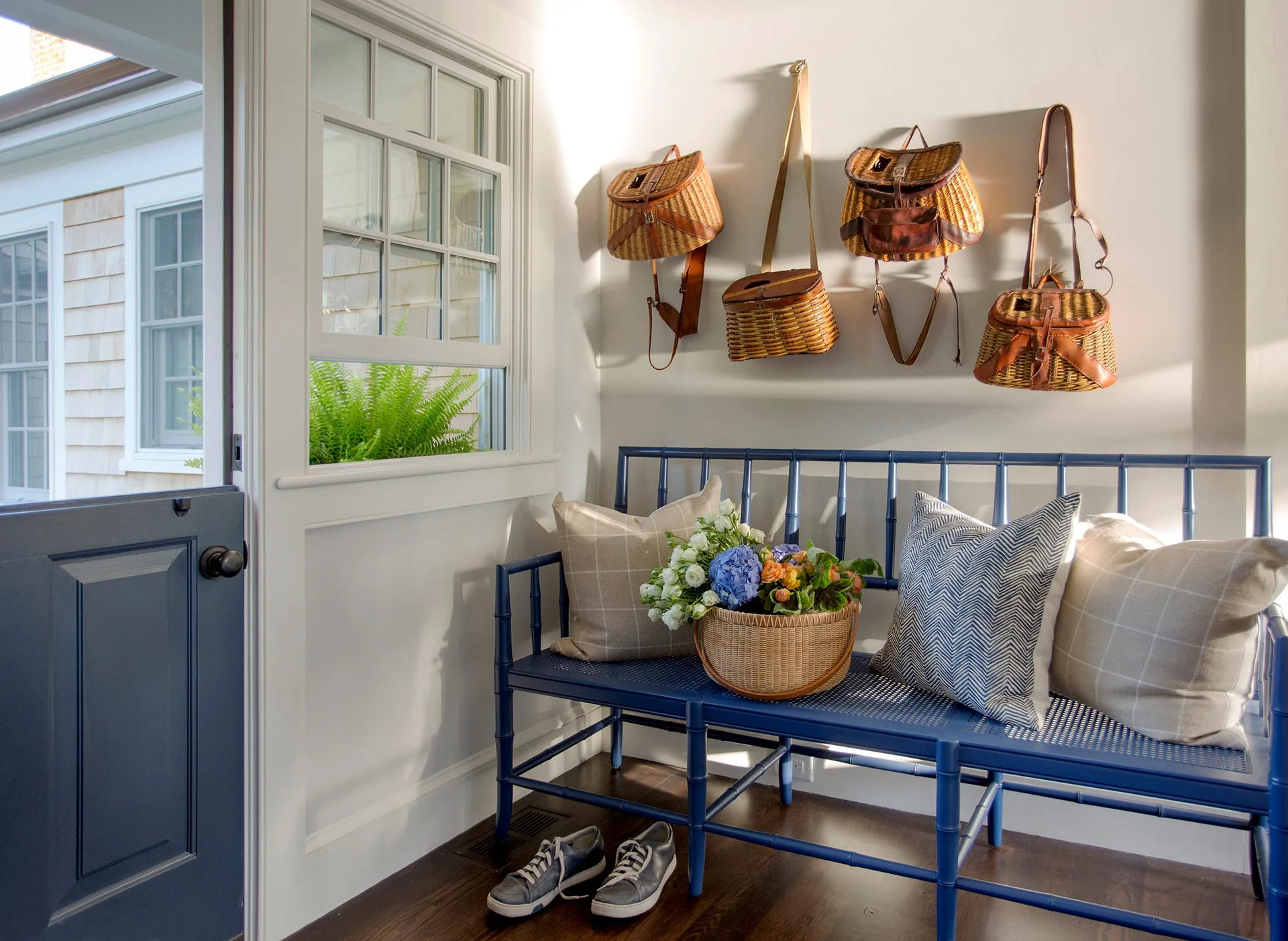 A blue bamboo-style bench with patterned pillows sits in a bright entryway next to a matching blue door and several hanging wicker baskets.