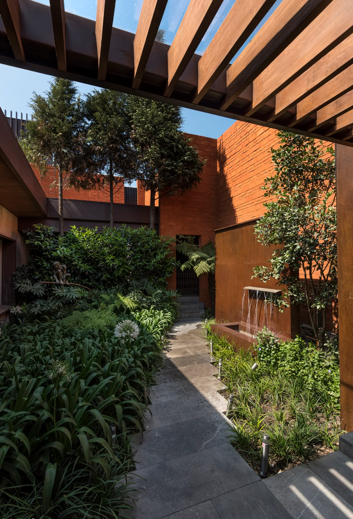 A stone pathway leads through a lush courtyard garden enclosed by red brick walls with a weathered metal water feature and overhead wooden pergola.