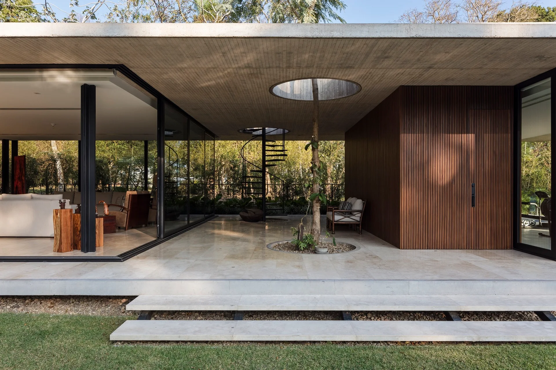 A covered modernist concrete patio featuring large stone floor tiles, a vertical wood slat wall, and a living tree growing through a roof cutout.