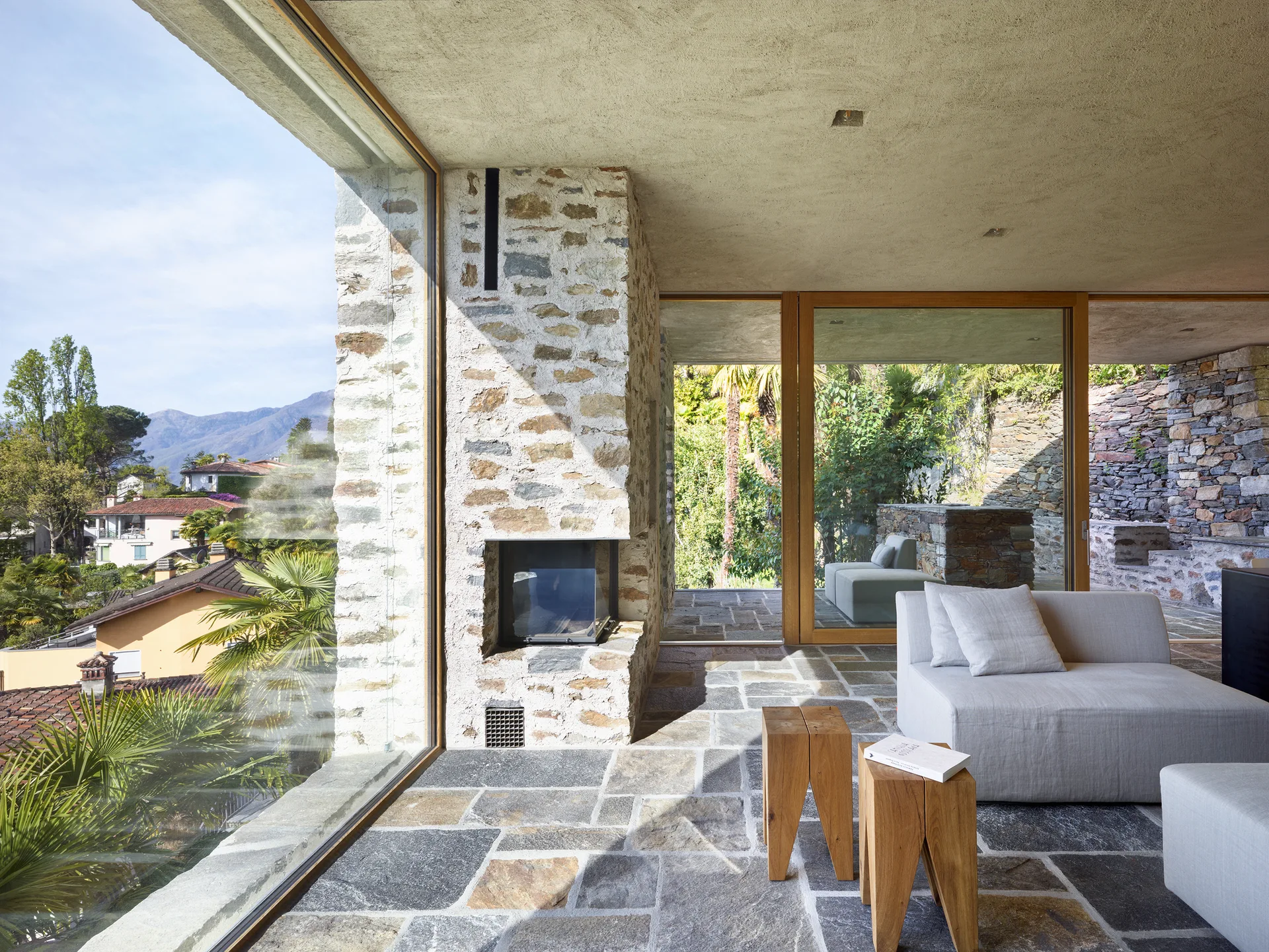 A modern minimalist living room features a rough stone fireplace, flagstone flooring, two wooden stool side tables, and floor-to-ceiling glass windows.