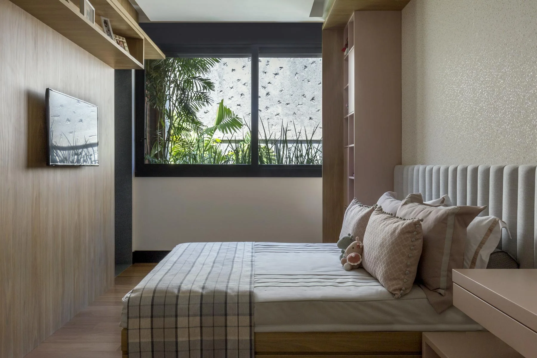 A modern bedroom features a bed with an upholstered headboard, integrated wood wall paneling with a television, and built-in shelving next to a window.