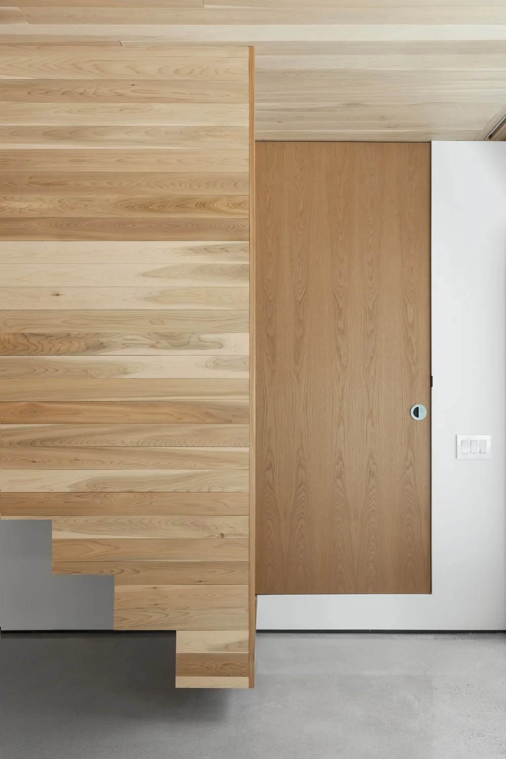 A minimalist interior wall featuring horizontal light wood paneling with a stepped profile next to a flush vertical grain wood door on a white wall.