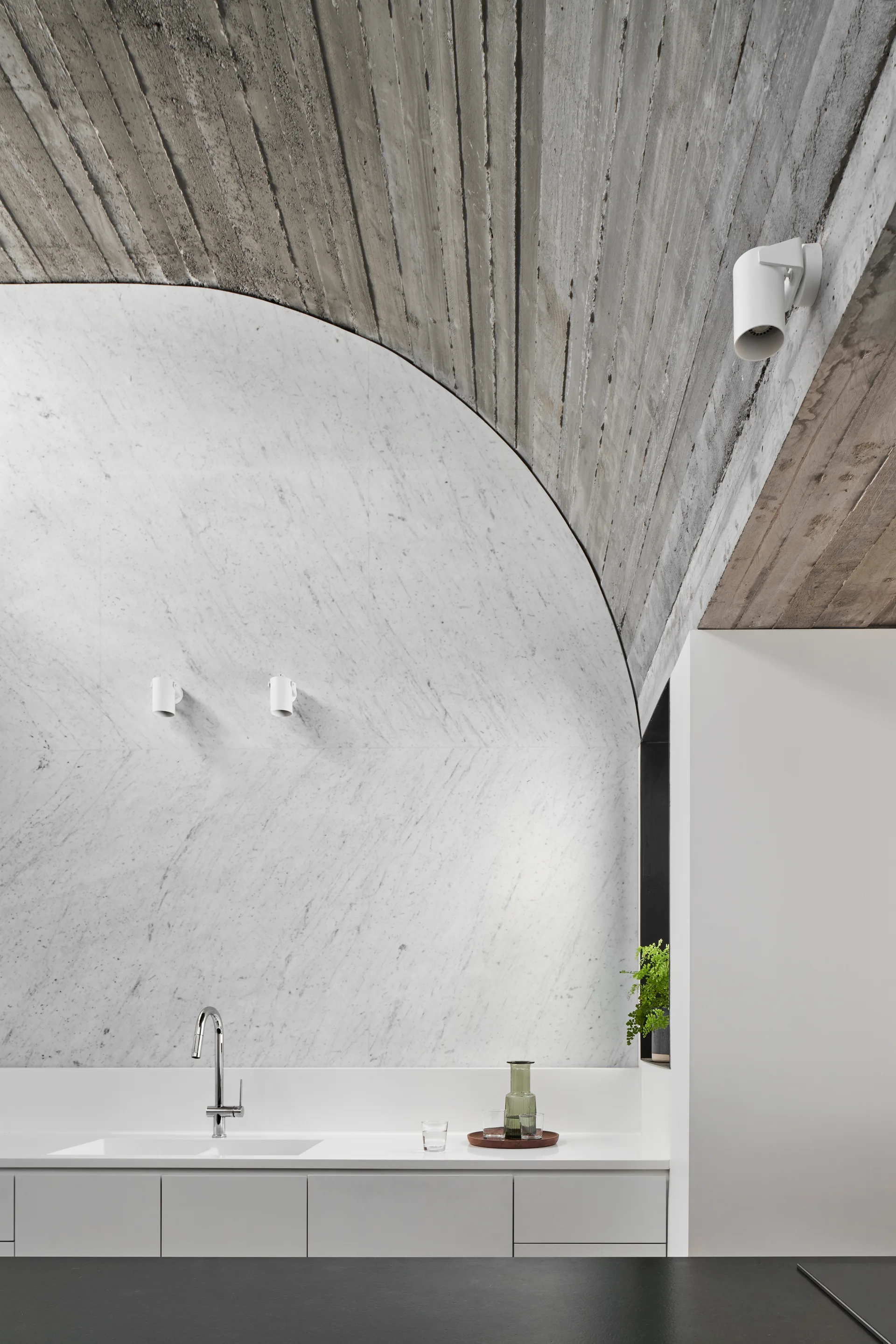 A minimalist kitchen counter with an integrated sink beneath a white marble arched wall and a textured, vaulted concrete ceiling.