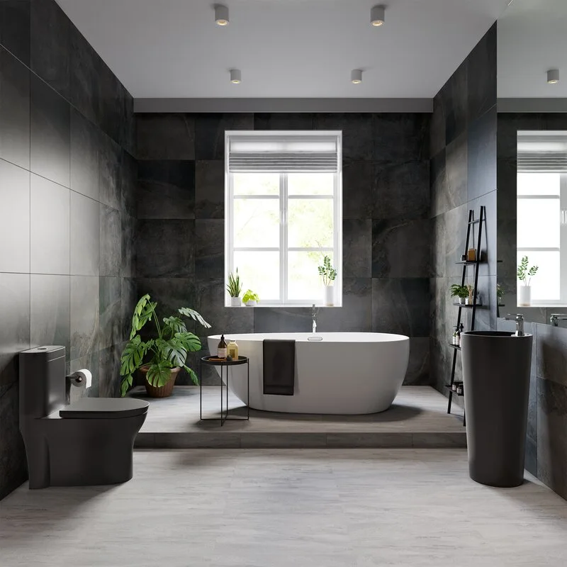 A modern bathroom featuring a white freestanding bathtub on a raised platform against charcoal grey tiled walls with a black toilet and pedestal sink.