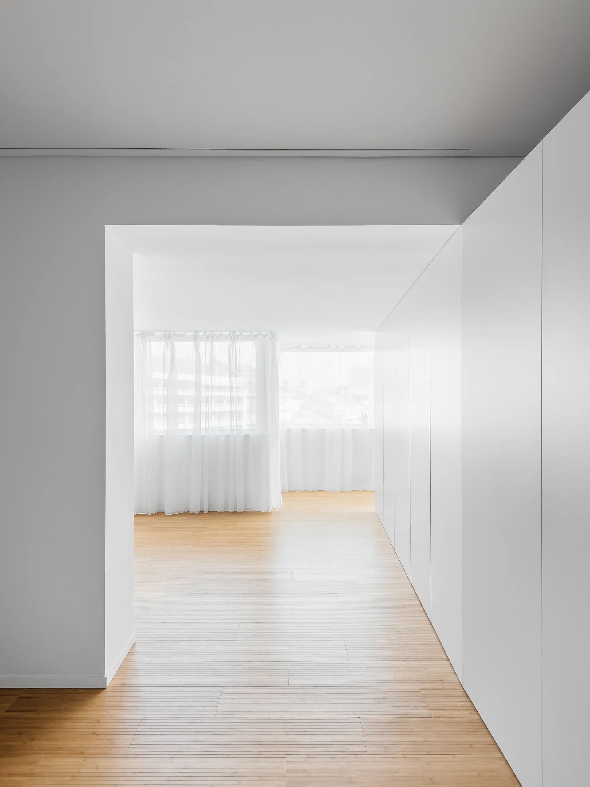 A minimalist hallway features seamless white floor-to-ceiling cabinetry, light wood flooring, and sheer white curtains filtering natural light.