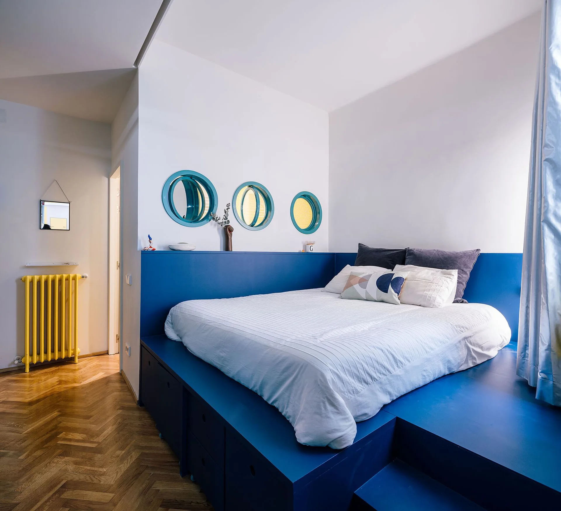 A bedroom with a custom blue storage platform bed, white walls featuring three round interior windows, and herringbone wood floors.