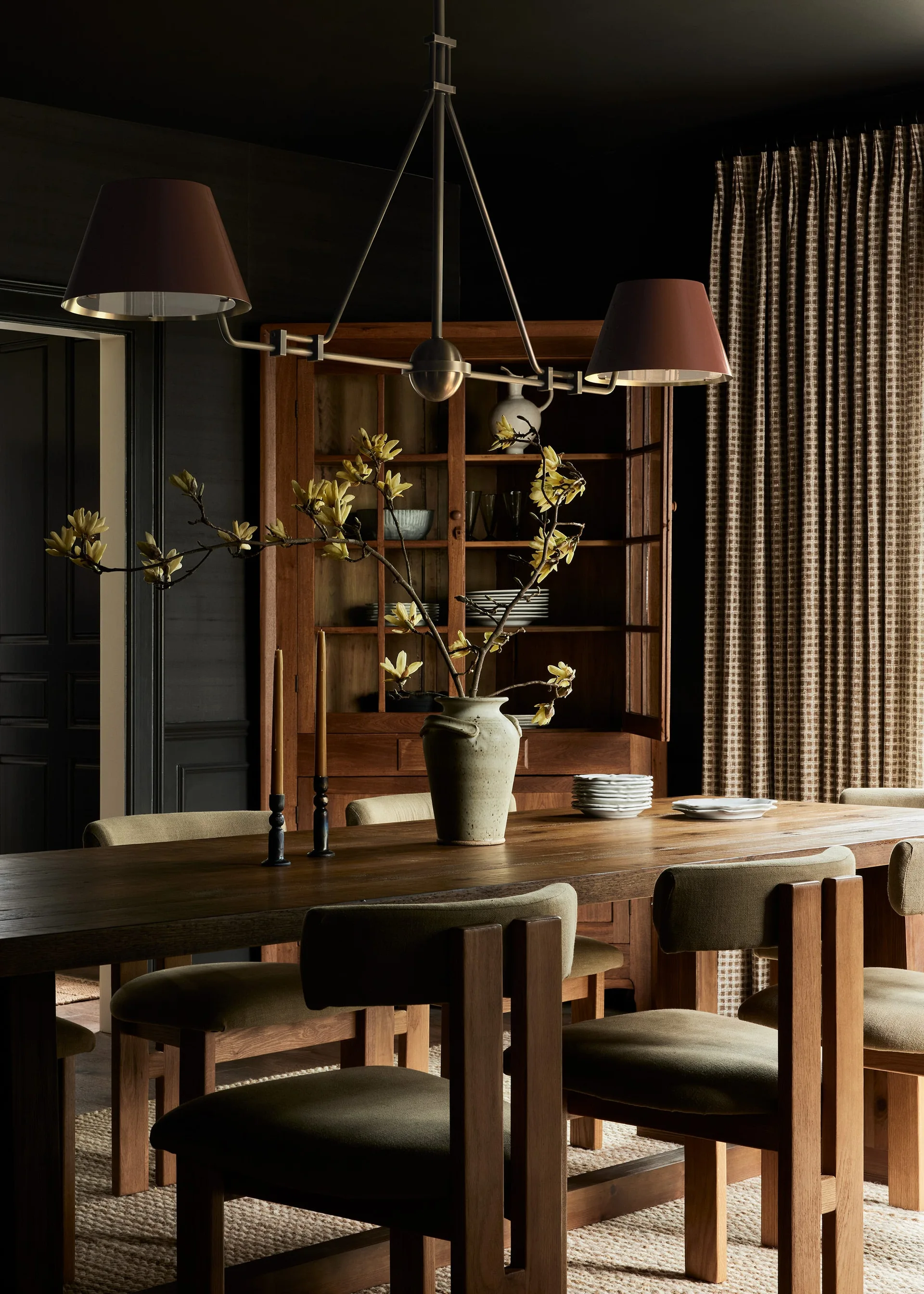 A dark dining room features a wooden dining table and chairs, a tall glass-front cabinet, and a ceramic vase with tall flowering branches.