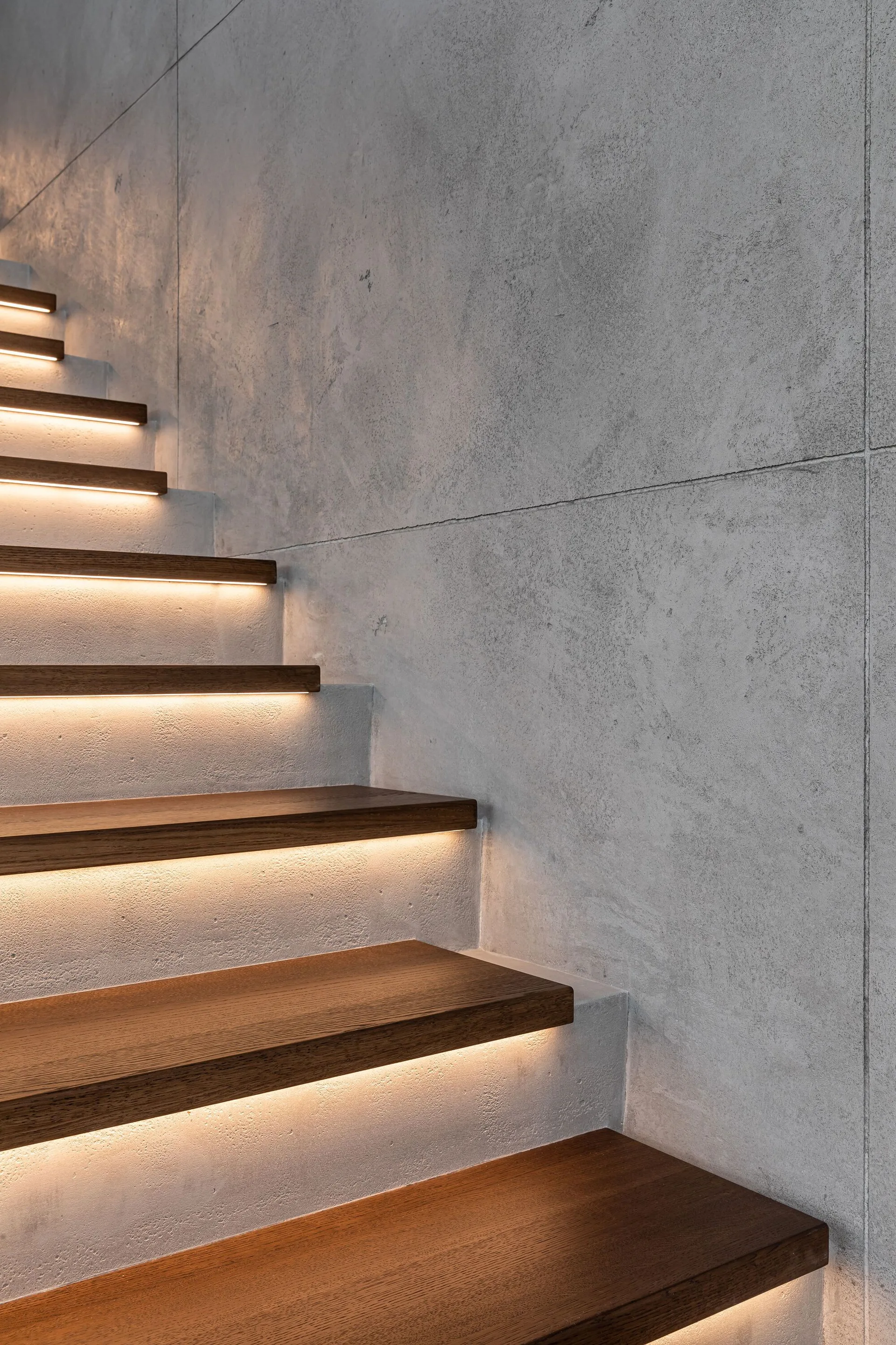 A close-up of a modern staircase featuring dark wood treads with integrated warm LED lighting on each step against a textured grey concrete panel wall.